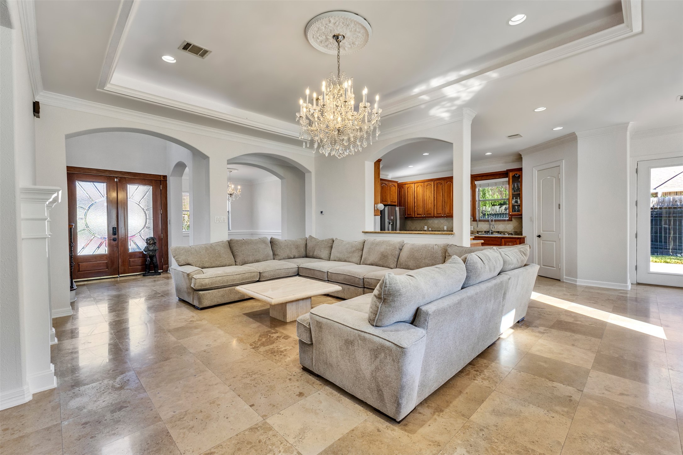 Elegant living room featuring high tray ceilings framed by intricate crown molding, a stunning chandelier with a decorative ceiling medallion, and graceful arched openings. Abundant natural light enhances the spacious, inviting atmosphere.
