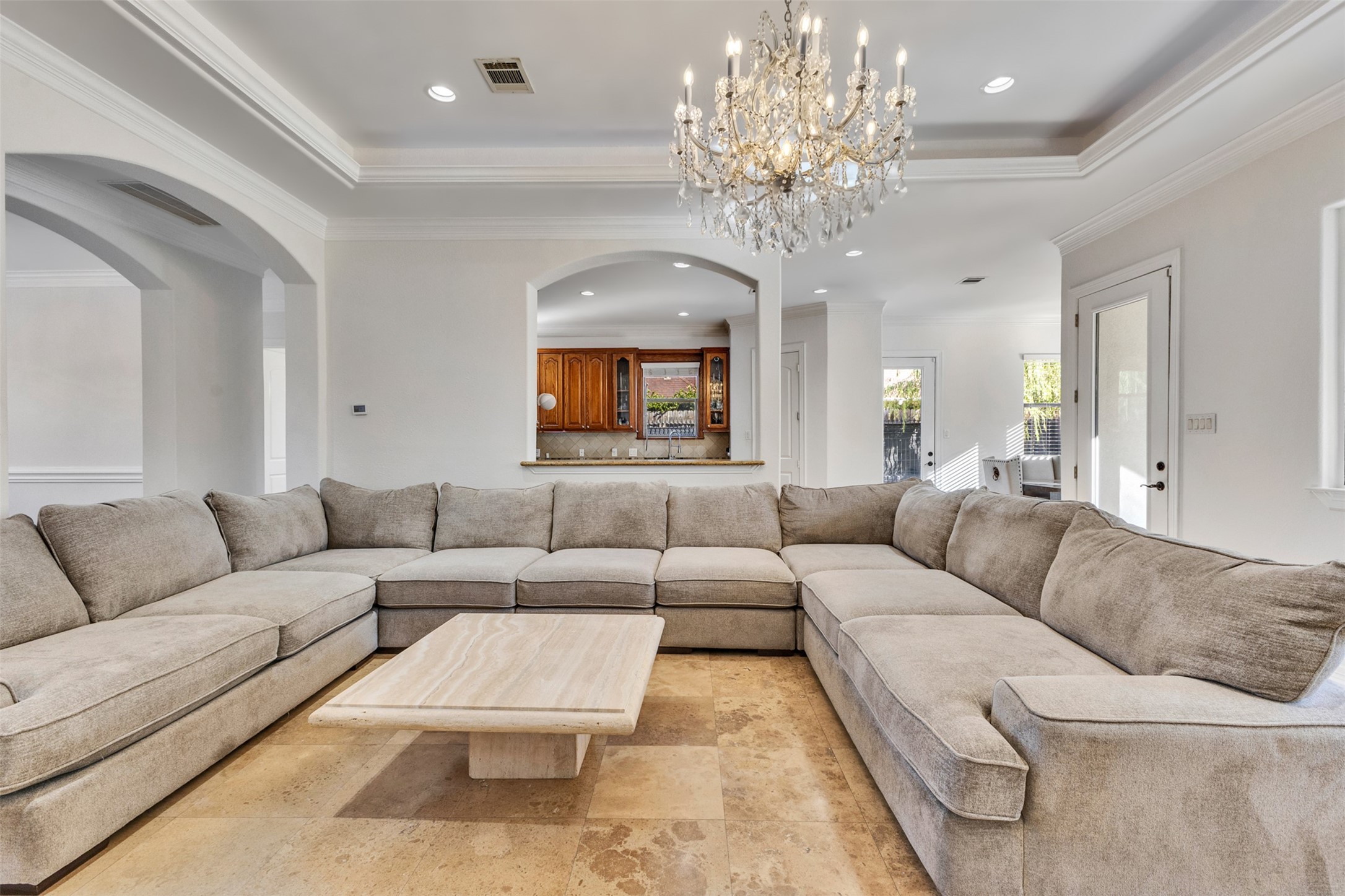 Elegant living room featuring high tray ceilings framed by intricate crown molding, a stunning chandelier with a decorative ceiling medallion, and graceful arched openings. Abundant natural light enhances the spacious, inviting atmosphere.