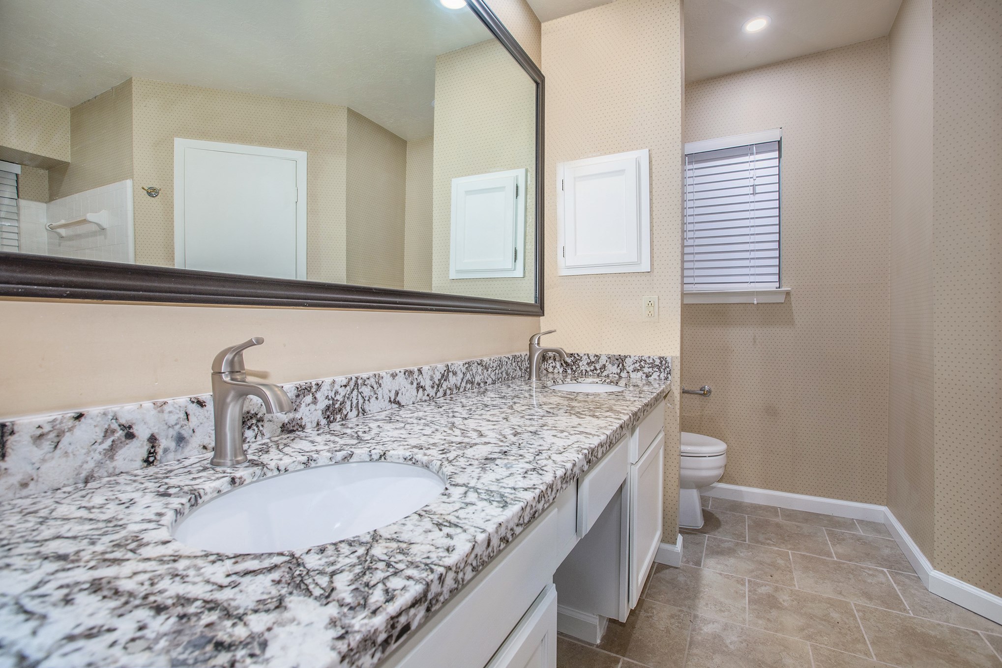 Master bathroom features granite countertop with dual sinks and faucets, framed mirror, white cabinets and tile flooring.