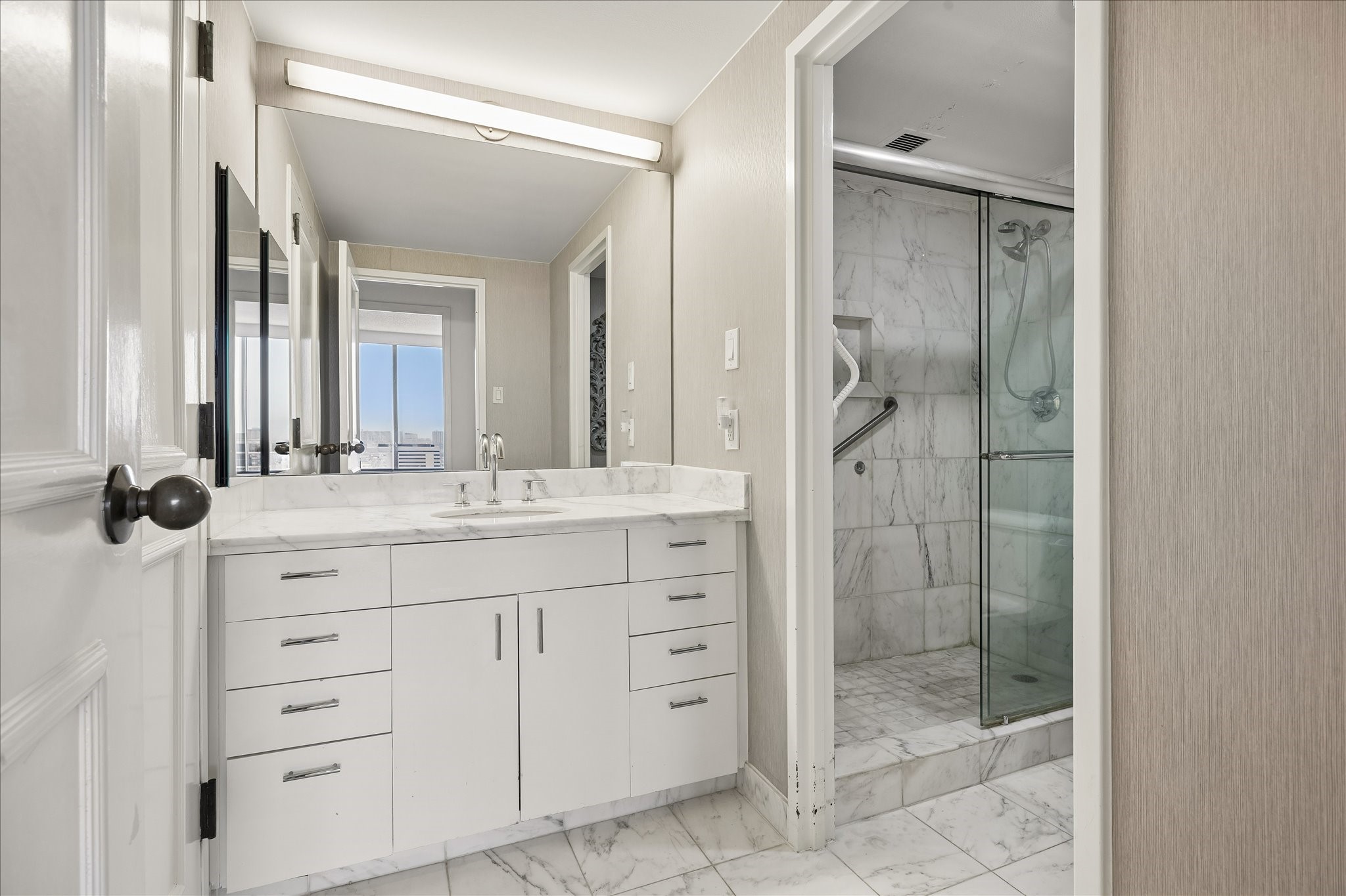 Drawer storage and a long vanity counter keep daily essentials organized. A large mirror and glass enclosed shower with light veined finishes complement the bathroom's practical layout.