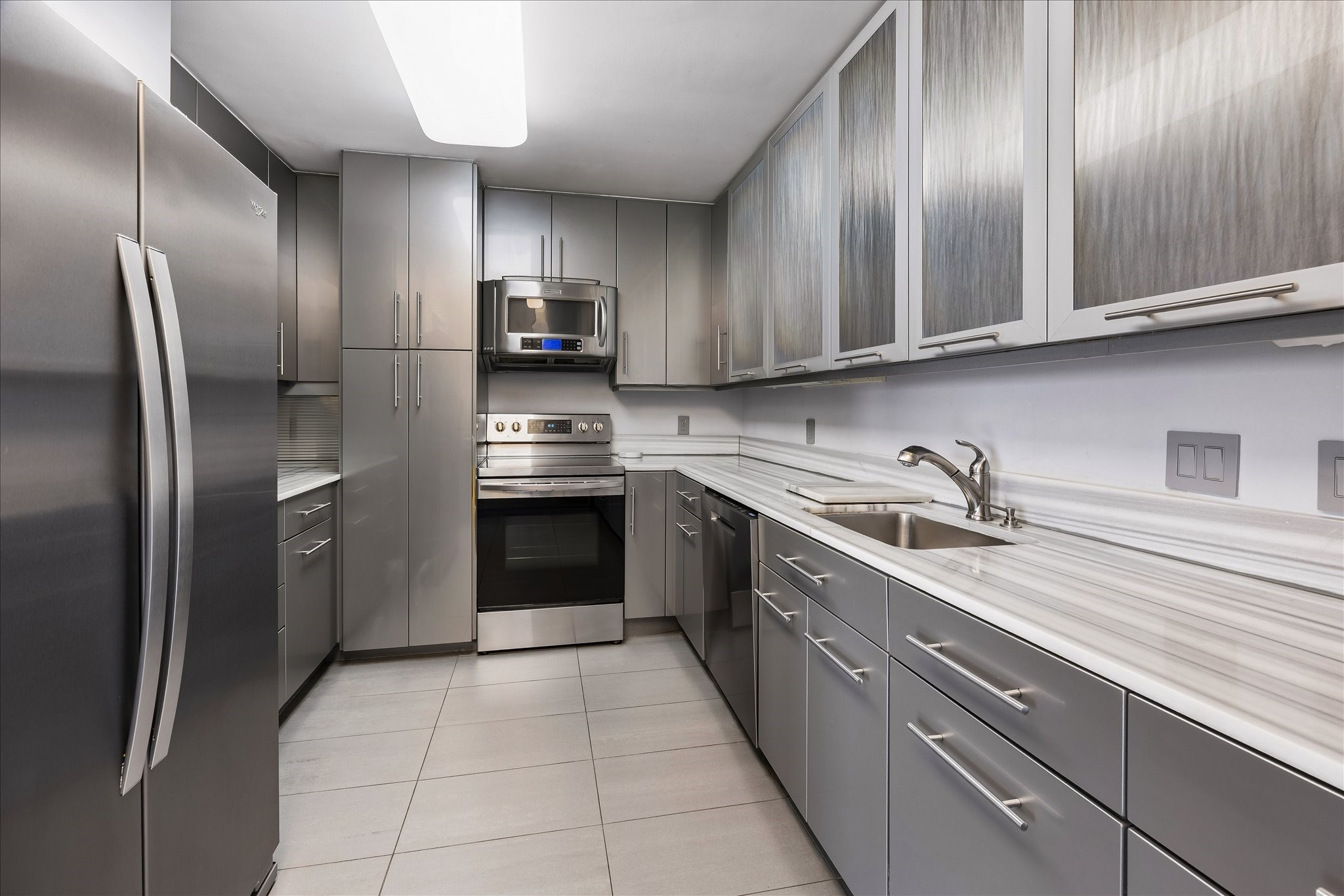 Gray cabinetry, textured glass upper doors and long counters define the galley kitchen. Storage and preparation areas line both sides, with stainless finish appliances supporting everyday cooking.