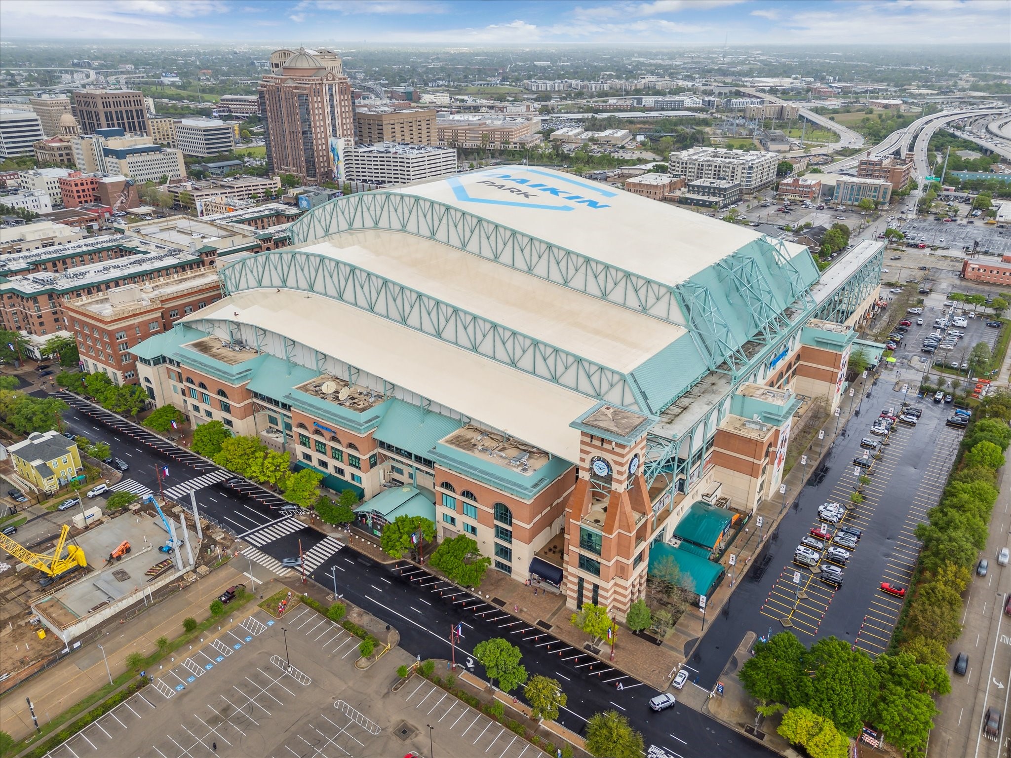 Soaring over downtown Houston, the photo captures Daikin Park, home of the Houston Astros, with its recognizable retractable roof and clock tower at the corner. Surrounding the ballpark, you can see a mix of modern office towers, historic brick buildings, and busy freeway interchanges stretching into the distance. The vibrant grid of city streets, parking areas, and tree-lined sidewalks emphasize its central location, while the open roofline stands out as a landmark in the skyline.