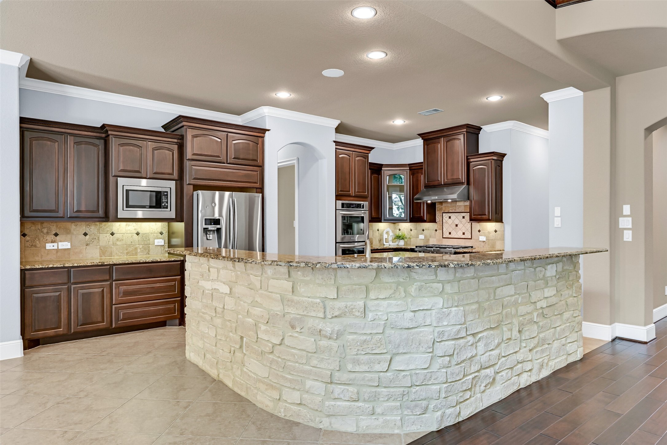 The island kitchen blends warmth and function with rich espresso cabinetry, granite countertops, undercabinet lighting, a travertine tile backsplash, and an oversized wraparound breakfast bar finished with stacked stone detailing that gives the space a custom feel.