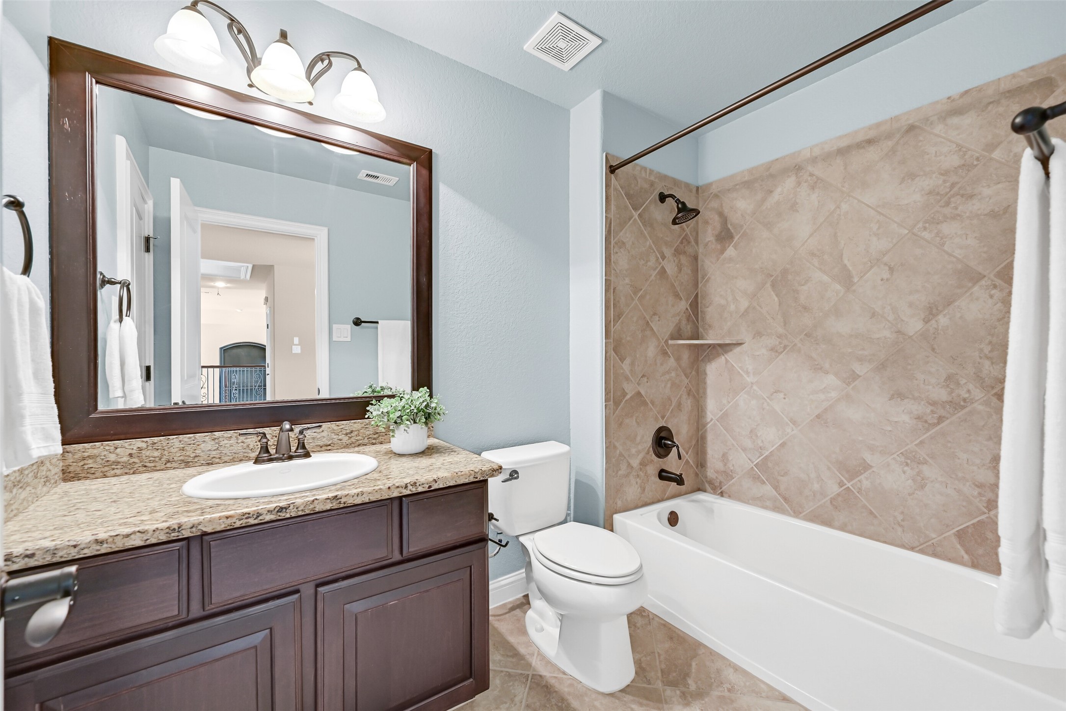 This private en suite bath features granite counters, a single basin sink, a framed mirror, tile flooring, and a beautifully tiled tub/shower surround with coordinating bronze fixtures.