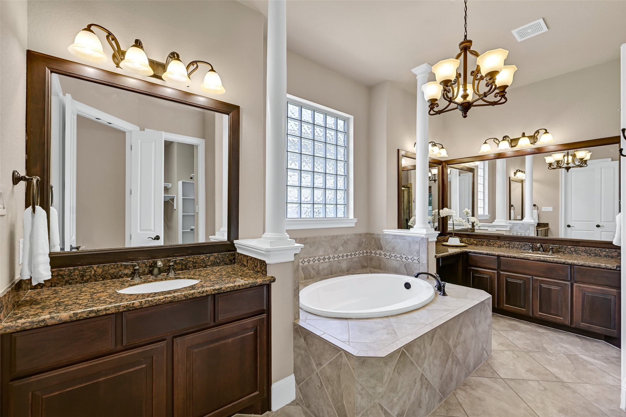 The spa-inspired primary bathroom features separate granite-topped vanities with rich espresso cabinetry, a soaking tub framed by decorative tilework and glass block windows, elegant lighting, and soaring ceilings that make the entire space feel open and luxurious.