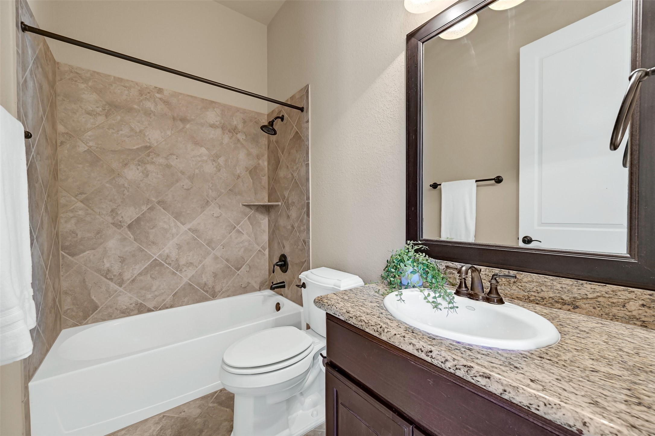 The en suite guest bath is finished with granite countertops, a single basin sink, rich wood cabinetry, oil-rubbed bronze fixtures, a framed mirror, and a tiled tub/shower surround that gives the space a clean, upscale feel.