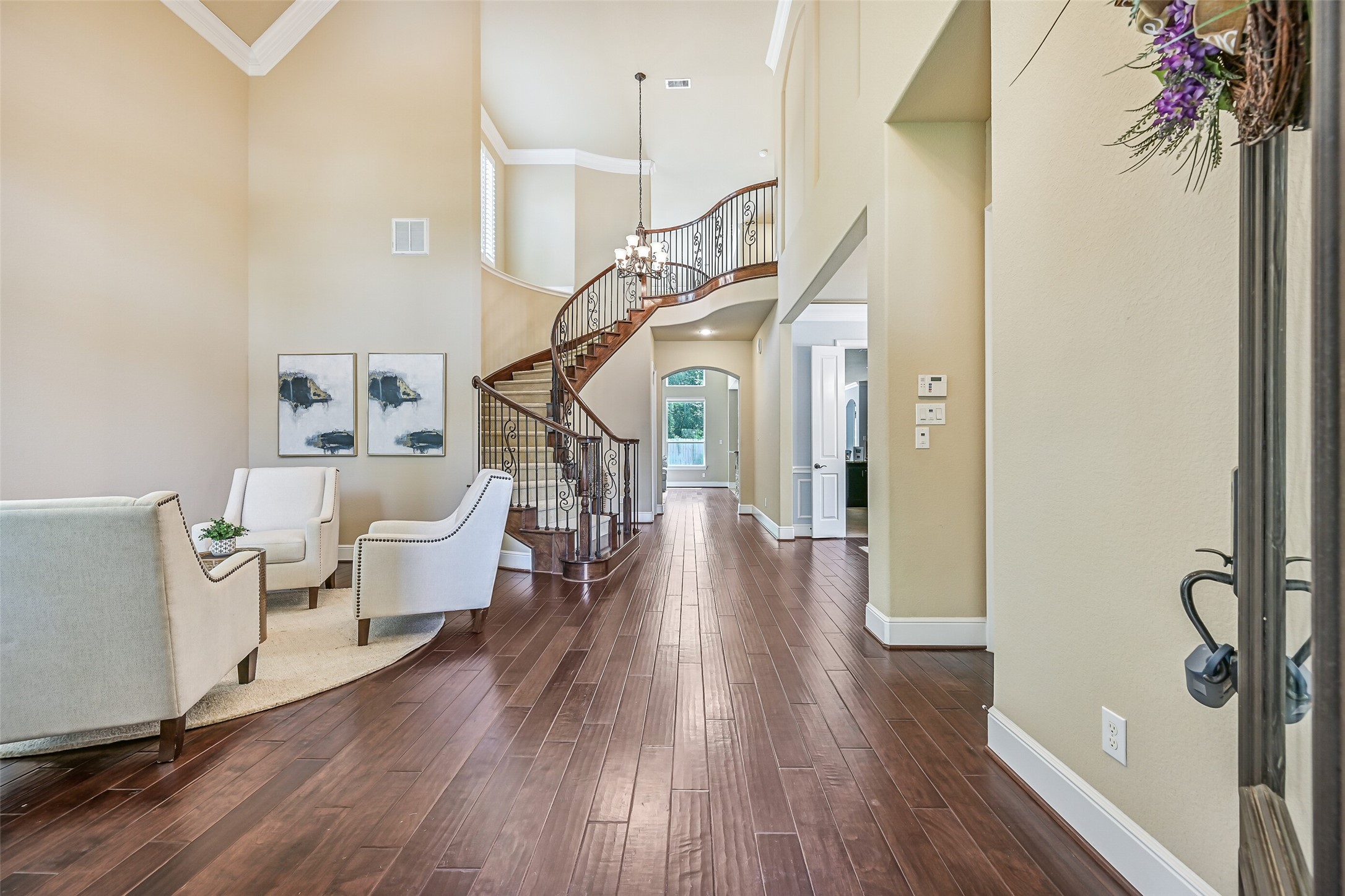 Step inside the grand foyer where soaring ceilings, engineered wood floors, and a dramatic curved staircase with iron balusters immediately set an elegant tone. A wall of towering windows trimmed with plantation shutters fills the space with natural light while highlighting the homes impressive architectural scale.