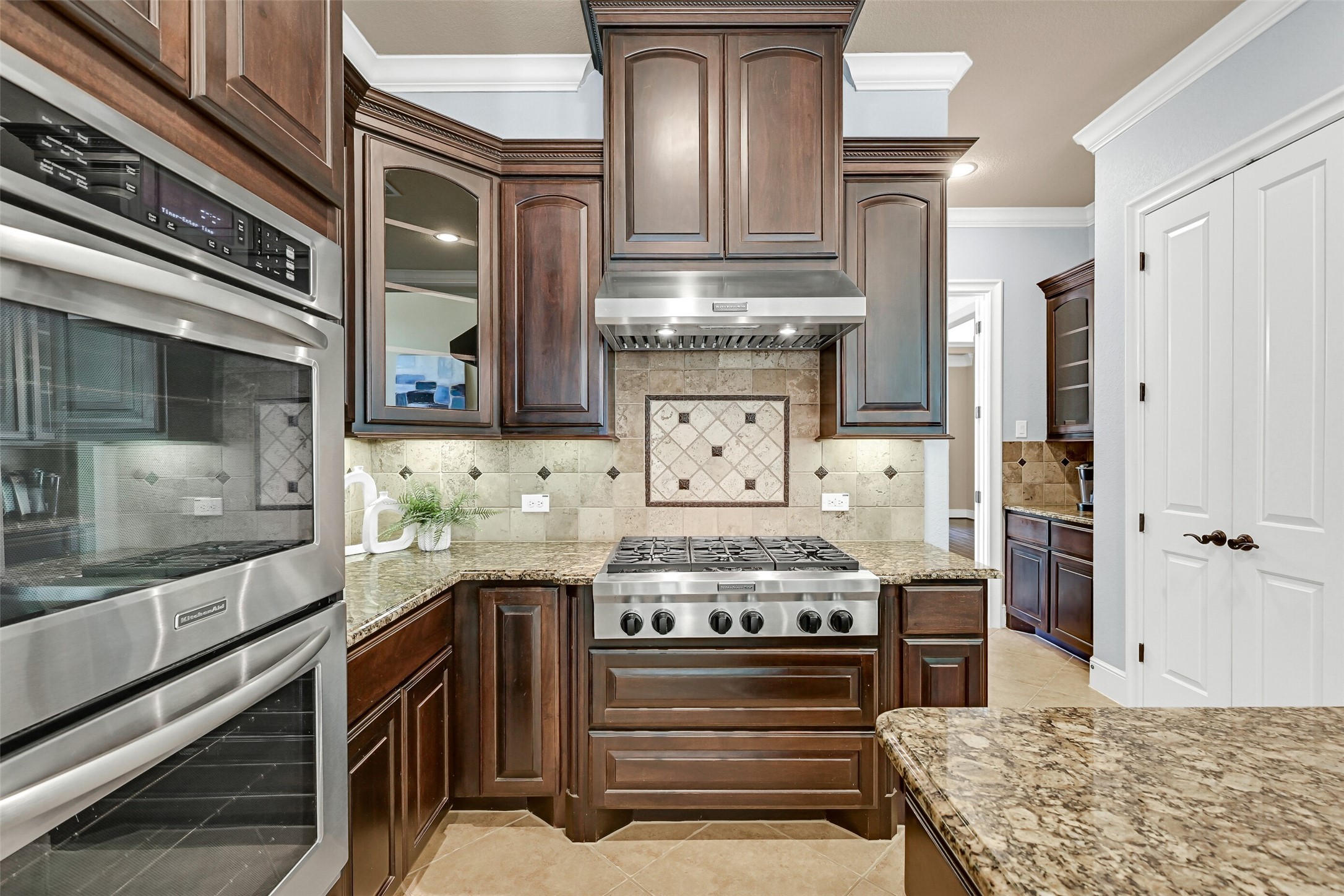 Designed for serious cooking, the kitchen features a six-burner KitchenAid gas cooktop beneath a vent hood framed by custom cabinetry and travertine backsplash details, along with dual electric convection ovens and abundant storage throughout.