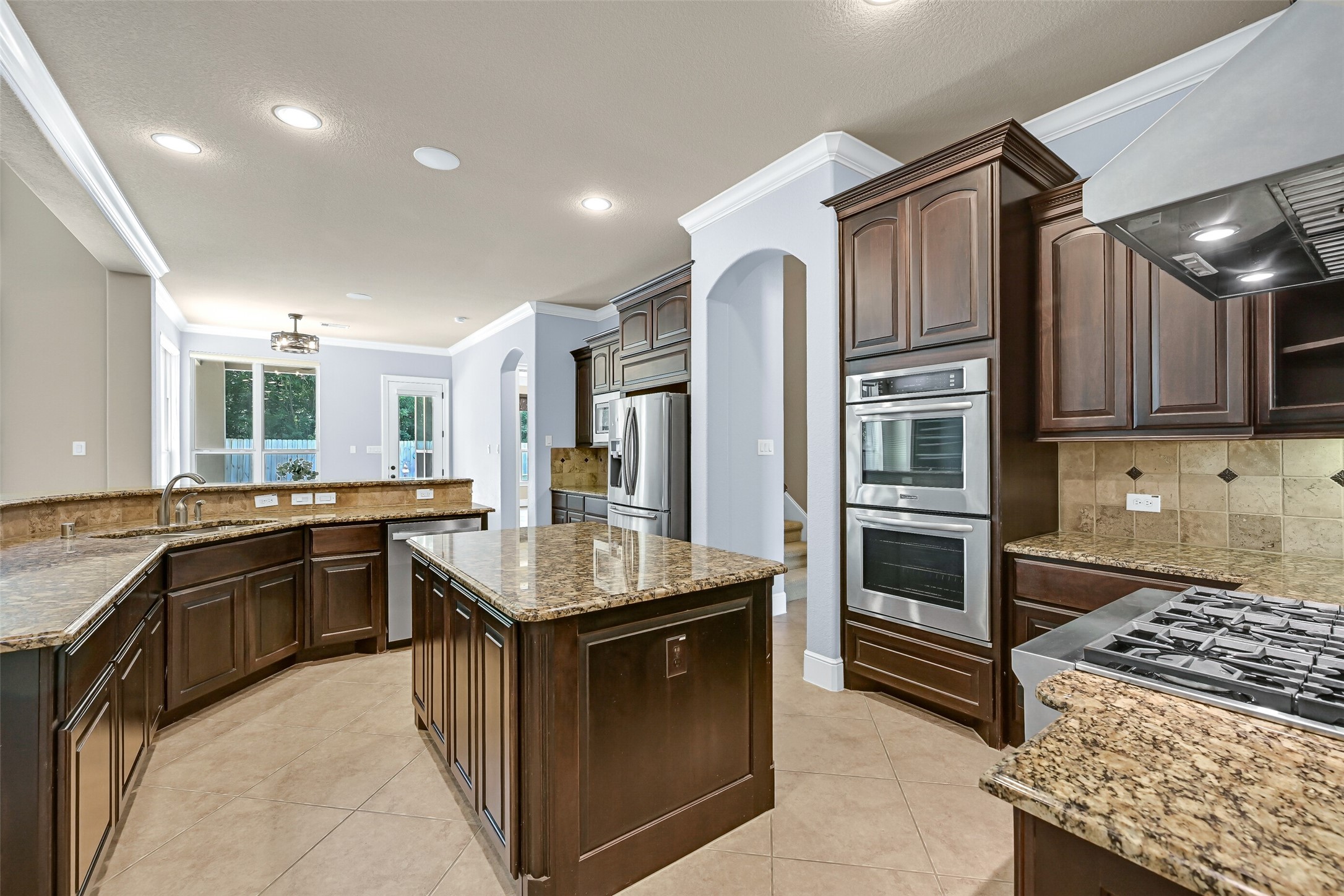 Another angle of the kitchen showcases the oversized island, extensive granite workspace, stainless appliances, and thoughtful layout that keeps everything within easy reach while still maintaining an open, airy feel. Through the arched entrance on right, you can catch a glimpse of the rear staircase of the home.