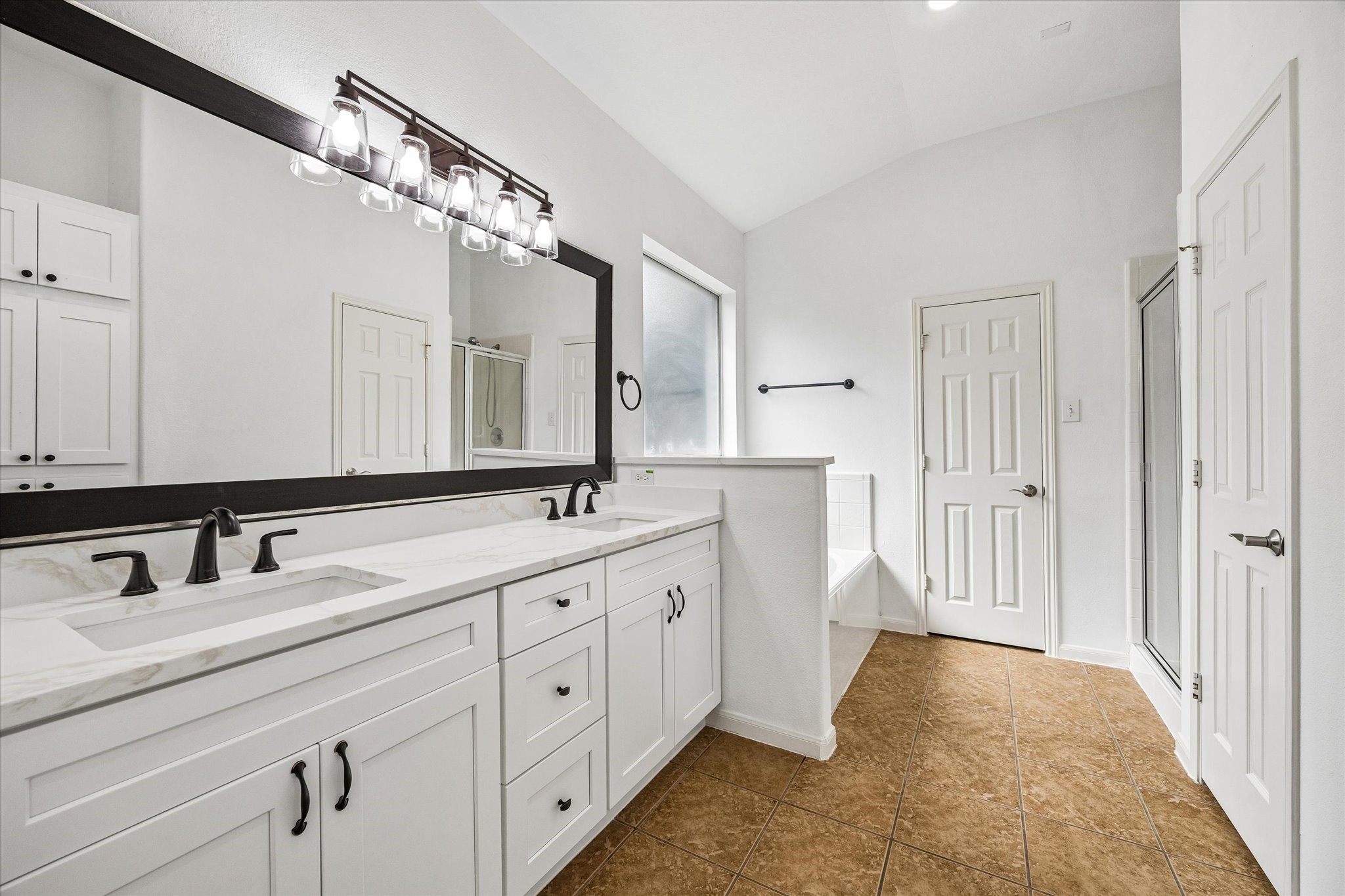 Spacious bathroom with a double vanity, sleek black fixtures, and a large mirror. Natural light flows in through the window, illuminating the elegant tile flooring and providing a bright, airy atmosphere.