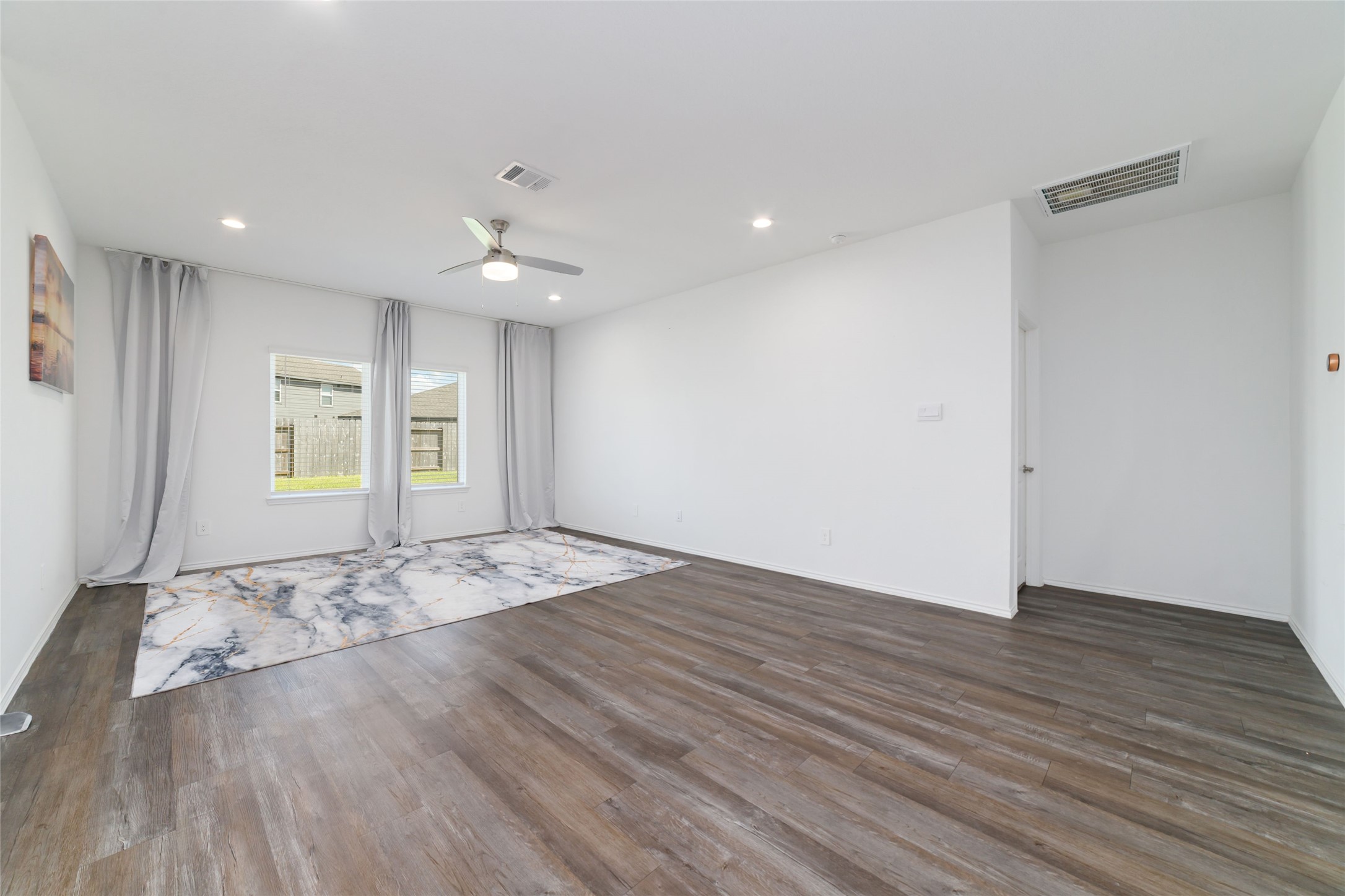 (Main Living Room): Sprawling, light-filled family room designed for effortless entertaining and relaxation. The space boasts continuous dark wood-look flooring, crisp white walls, recessed lighting, and a modern ceiling fan, while twin double windows framed by custom floor-to-ceiling drapery offer peaceful, unobstructed views of the private backyard.