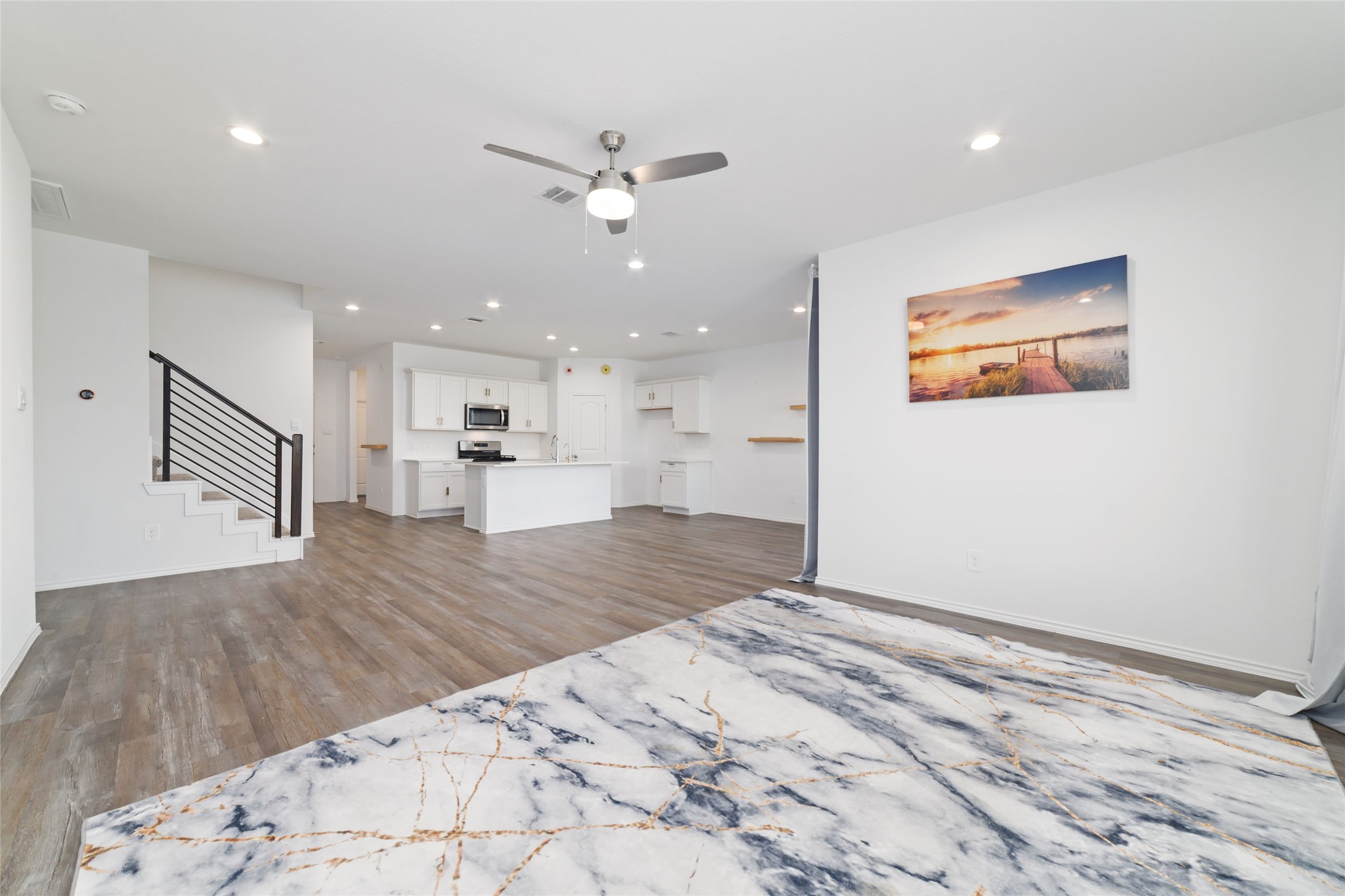 Sprawling open-concept kitchen and living area featuring dark wood-look flooring, a large central island with a solid countertop, white cabinetry, stainless steel appliances, and a carpeted staircase leading to the upper level