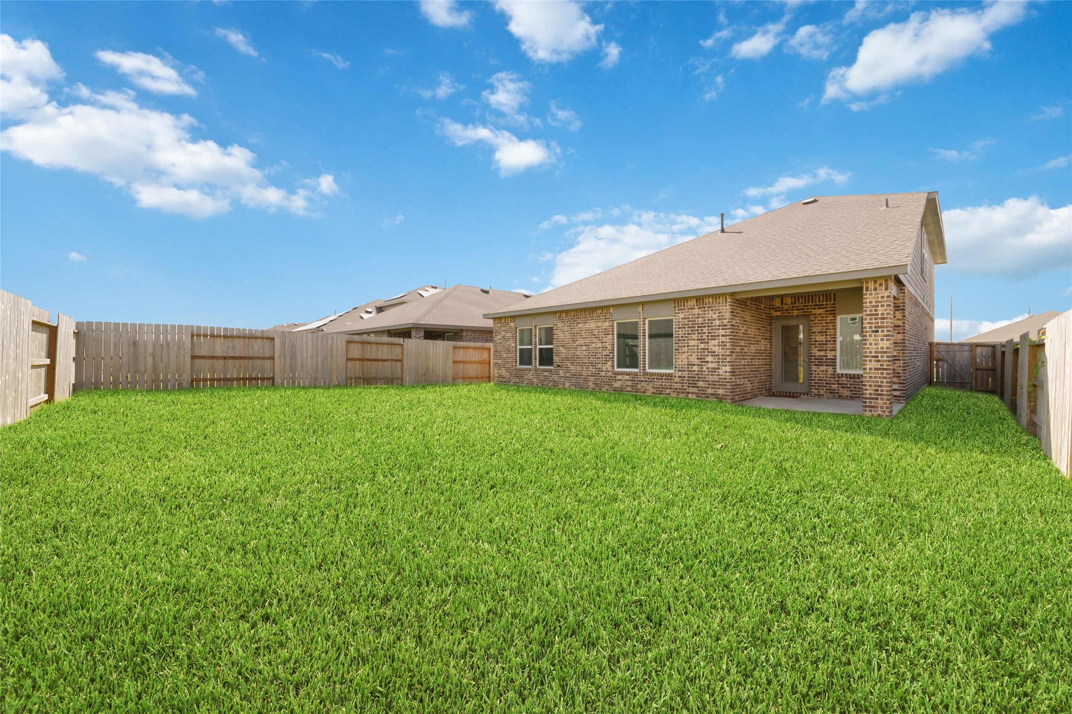 Full backyard perspective showing the large, manicured lawn completely enclosed by a tall wooden privacy fence, leading to a covered concrete patio attached to the rear of the brick home