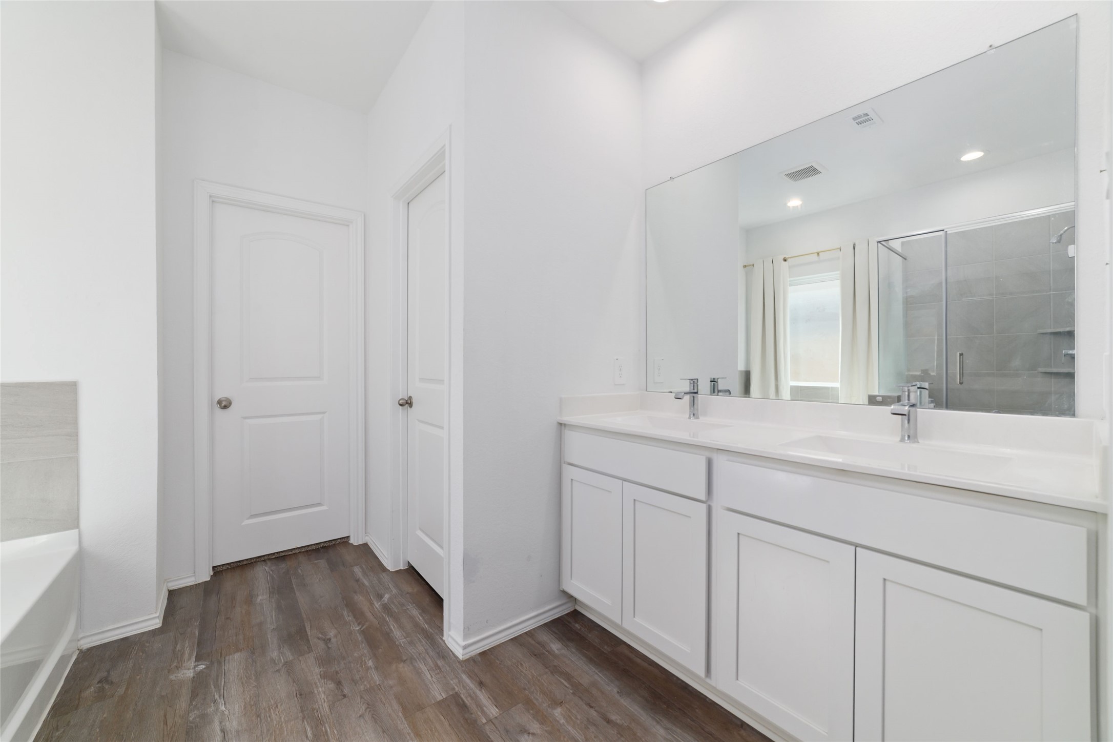 Interior view of a modern full bathroom featuring dark wood-look flooring, a white vanity with an undermount sink, a large mirror, and a tiled shower/bathtub combination with standard fixtures