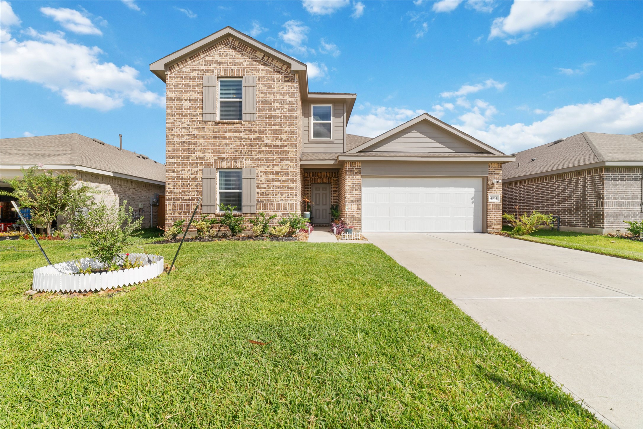 Exterior front view of a newly-constructed two-story house with a brick facade, contrasting shutters, a broad concrete driveway, a green lawn with a newly-planted tree, under a bright blue sky Exterior front view of a newly-constructed two-story house with a brick facade, contrasting shutters, a broad concrete driveway, a green lawn with a newly-planted tree, under a bright blue sky