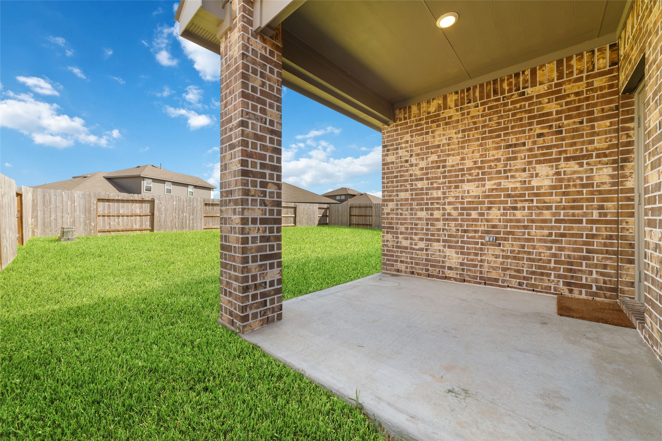 Invitation view of the covered concrete patio area, highlighting the robust brick pillar, simple mat, and the space for outdoor relaxation, looking out over the private lawn.