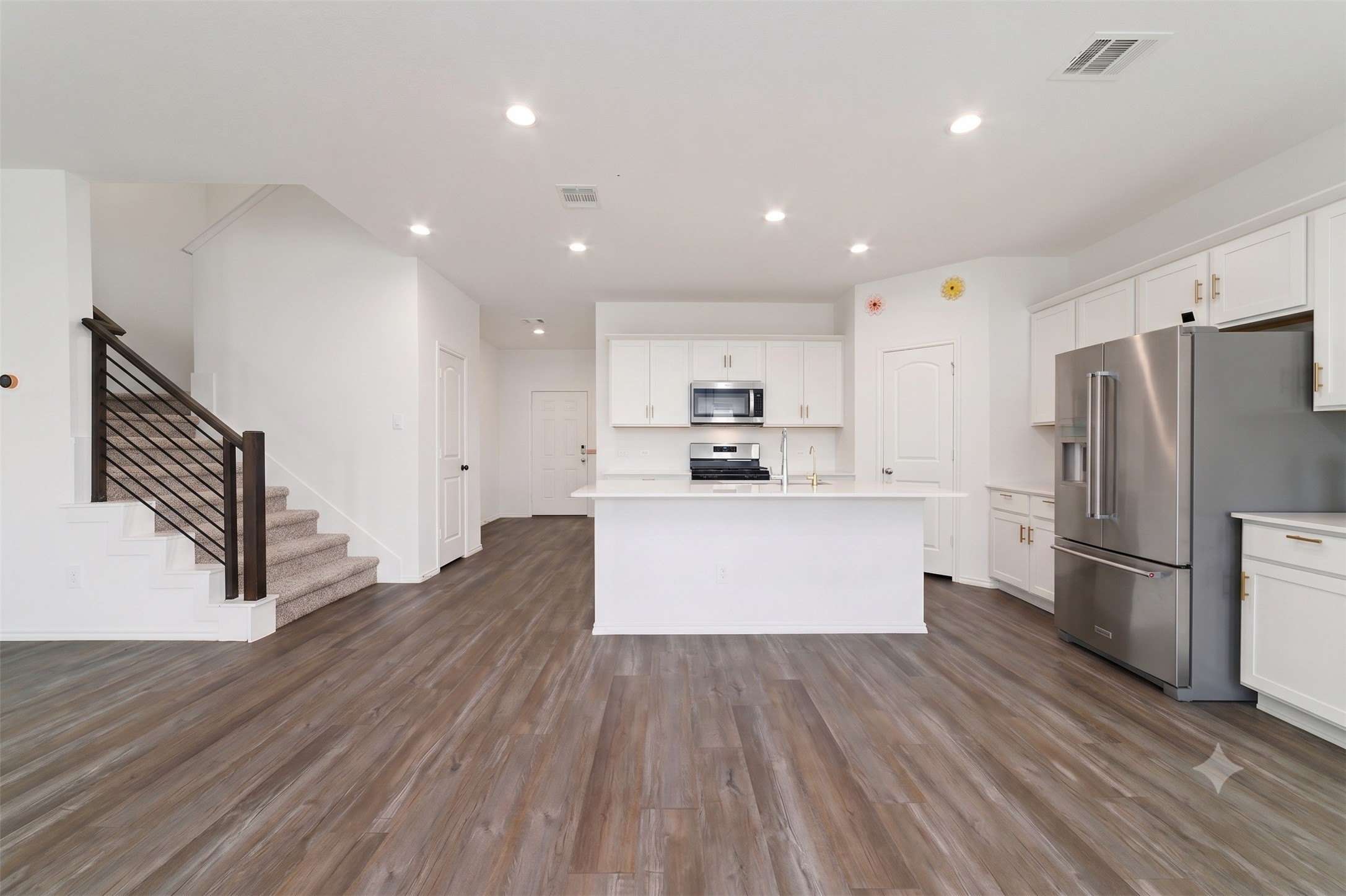 A complementary wide view of the modern open-plan kitchen and living space, showing the full appliance suite, central island, and the main staircase under recessed lighting. A complementary wide view of the modern open-plan kitchen and living space, showing the full appliance suite, central island, and the main staircase under recessed lighting.