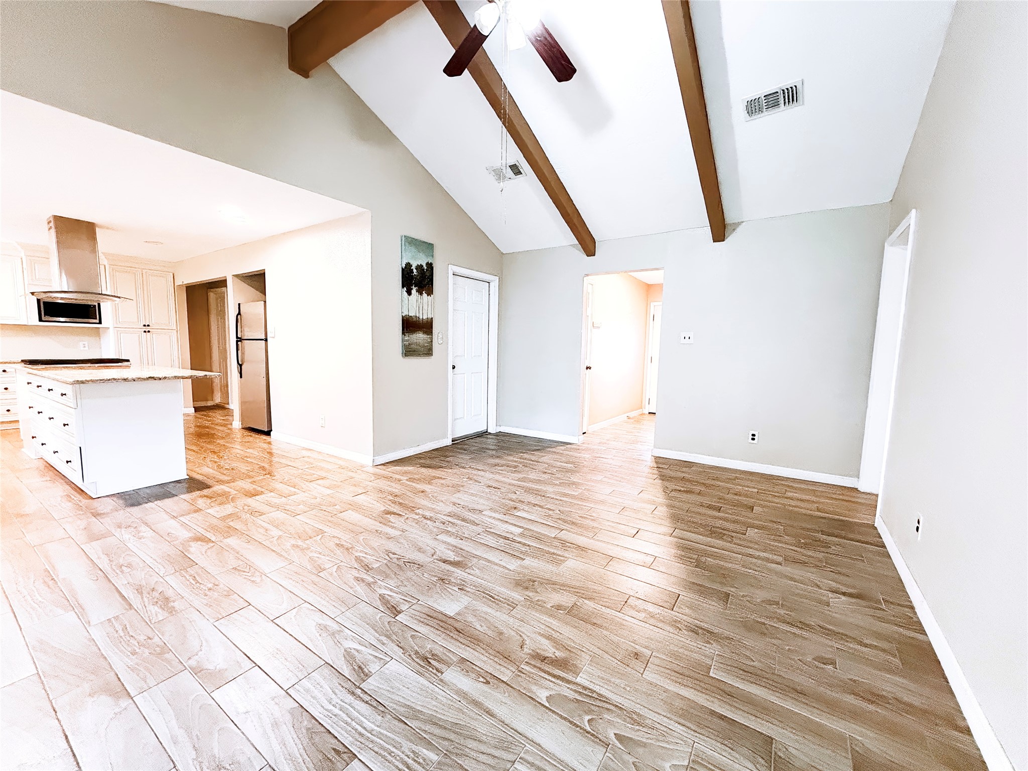 Spacious open-concept living area with vaulted ceilings, exposed wooden beams, and elegant wood-look tile flooring leading into a bright, modern kitchen.