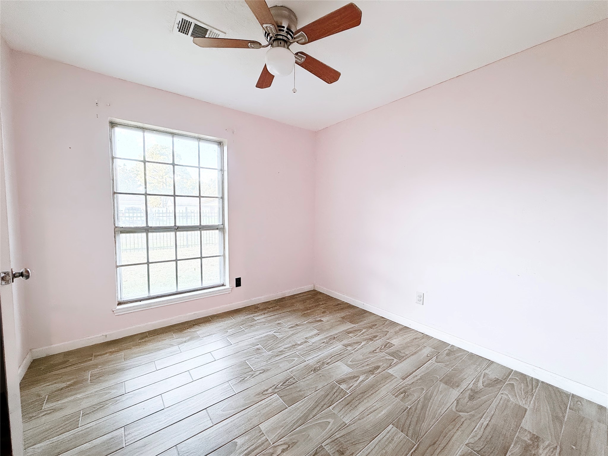 Charming bedroom with soft pink walls, wood-look tile flooring, and abundant natural light from a large window, complemented by a ceiling fan.