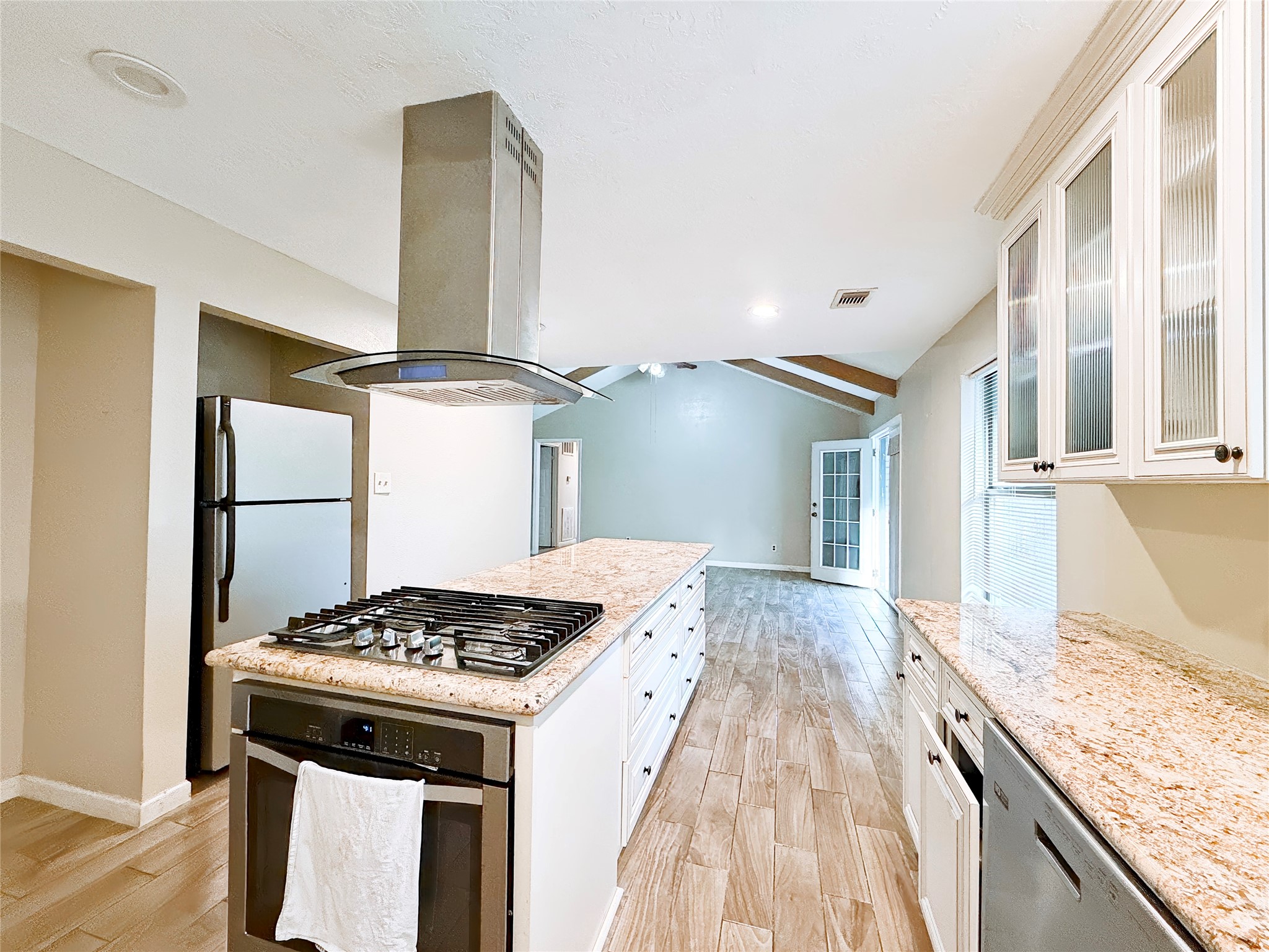 Modern kitchen with granite countertops, a central island featuring a gas cooktop, and stainless steel appliances. The open layout flows seamlessly into a bright living area with vaulted ceilings and wood-look flooring.