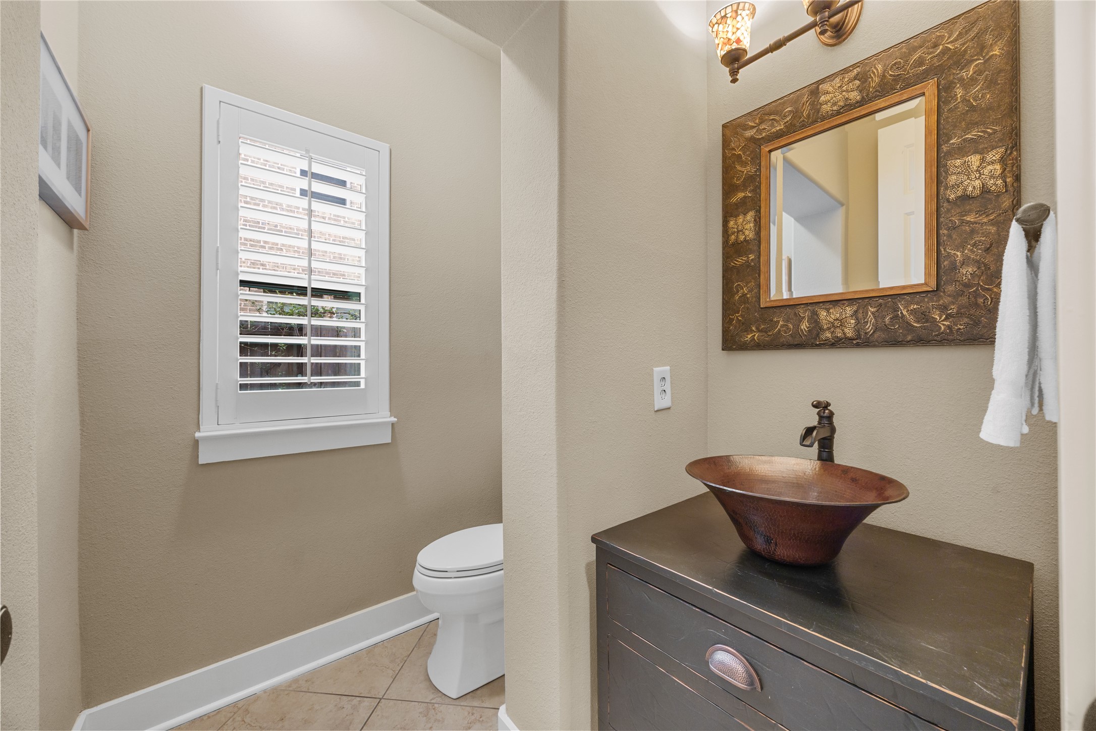 Charming powder room featuring a distinctive copper-toned vessel sink, decorative mirror, and plantation shutters.