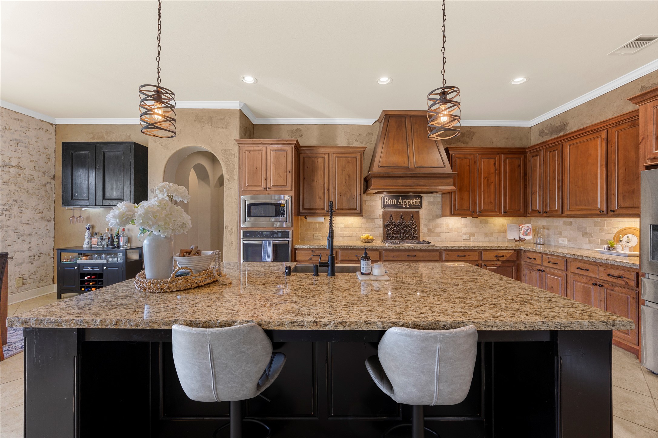 The kitchen's oversized granite island offers generous prep space and seating, complemented by rich wood cabinetry, pendant lighting, stainless steel appliances, and a custom vent hood.