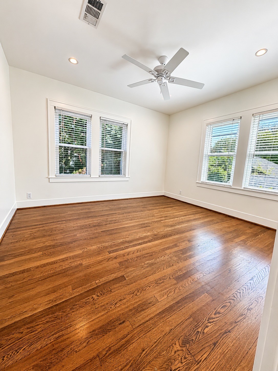 Generously sized bedroom with hardwood floors, ceiling fan, and natural light from windows on two walls.