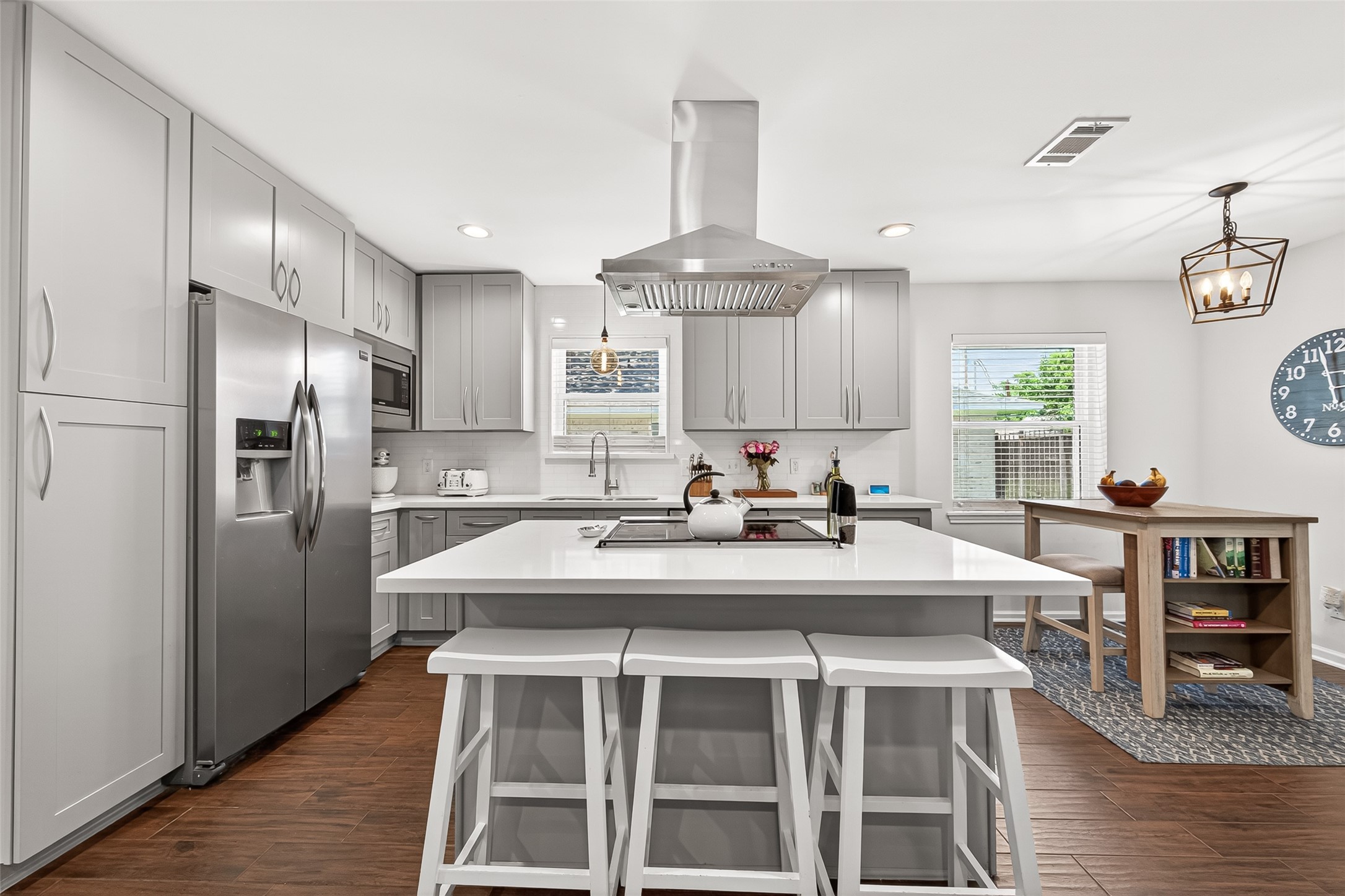 This modern kitchen features sleek gray cabinets, stainless steel appliances, and a central island with bar seating. It has ample natural light, wood-like tile flooring, and a cozy dining nook, creating a stylish and functional space.