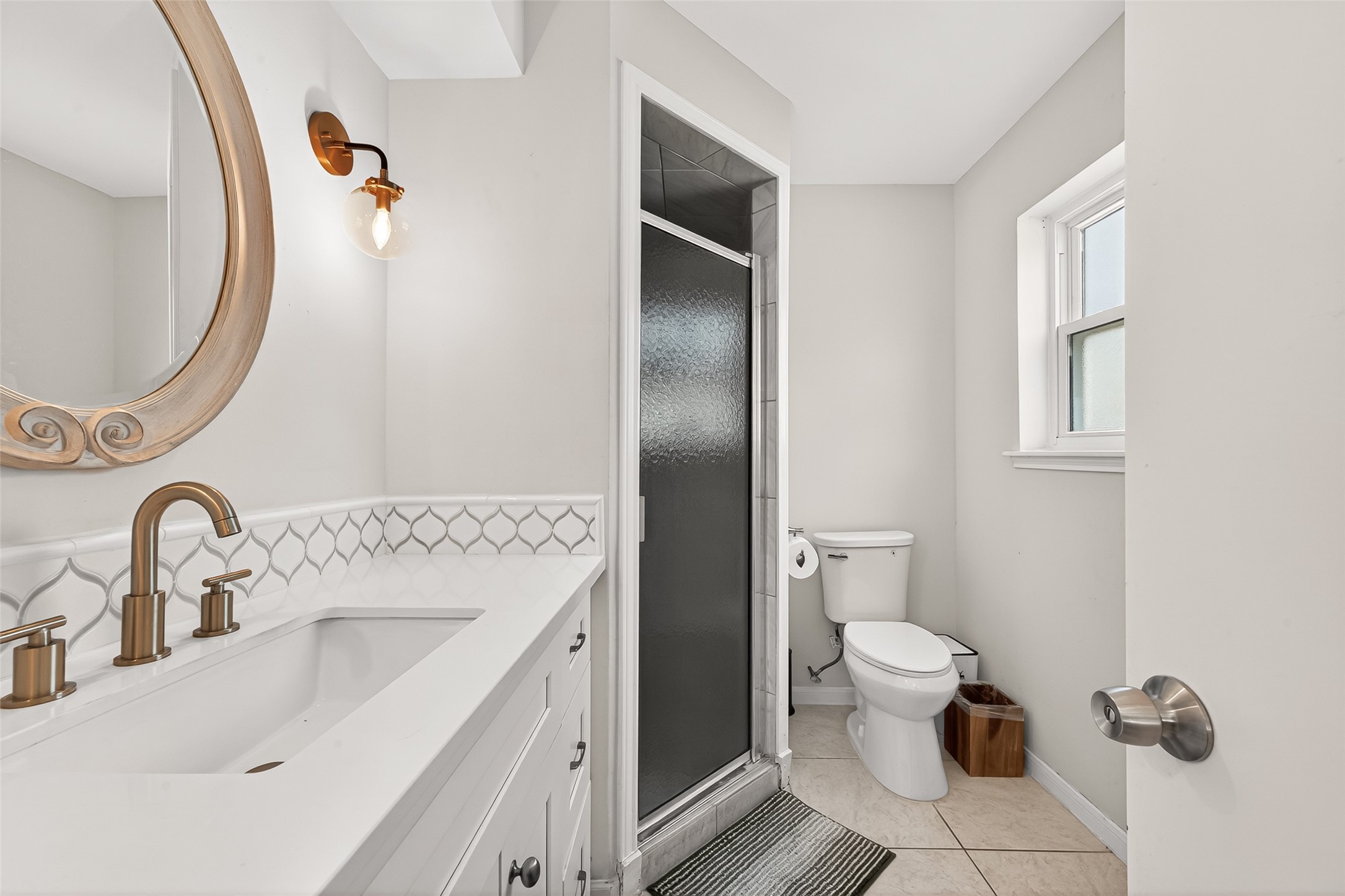 This primary bathroom features a modern design with a large mirrored vanity, elegant fixtures, and a sleek glass shower. Natural light illuminates the space through a window, creating a bright and welcoming atmosphere.