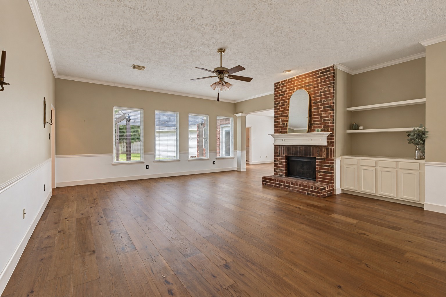 This large family room offers generous space for gathering and entertaining, with engineered wood flooring and a brick wood-burning fireplace as its centerpiece. Built-in shelving and cabinetry provide storage and display space, while crown molding and classic wainscoting add character. Four tall windows fill the room with natural light and offer views of the backyard. This large family room offers generous space for gathering and entertaining, with engineered wood flooring and a brick wood-burning fireplace as its centerpiece. Built-in shelving and cabinetry provide storage and display space, while crown molding and classic wainscoting add character. Four tall windows fill the room with natural light and offer views of the backyard.