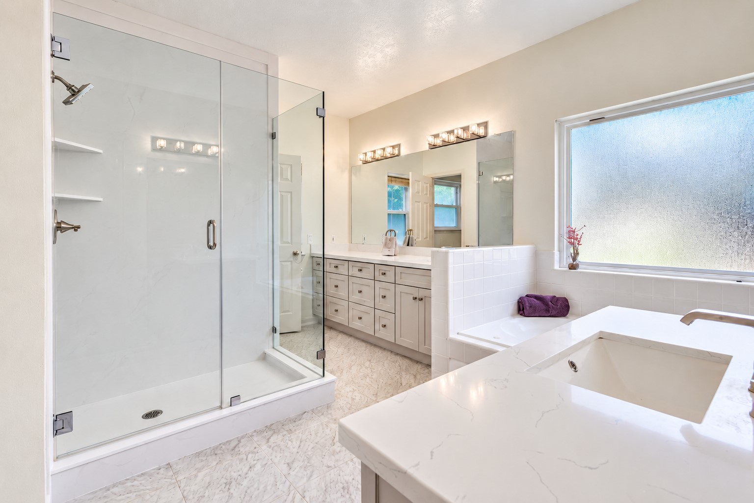 The spacious primary bath features a large shower with a frameless glass enclosure, a separate jetted tub, and dual vanities with ample cabinet and drawer storage. Light finishes and a large privacy window create a bright, inviting space to unwind.