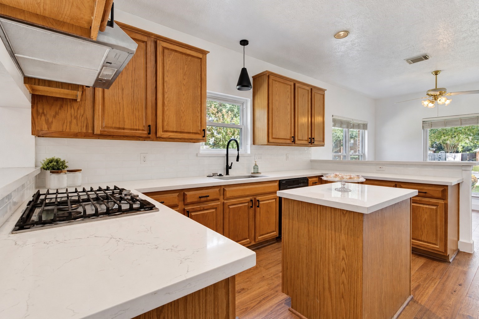 This inviting kitchen pairs warm wood cabinetry with quartz countertops, a crisp white subway tile backsplash, and contrasting black hardware. A gas cooktop, center island, and generous counter space make meal preparation easy, while abundant cabinetry provides ample storage. Natural light and an open connection to the breakfast area create a welcoming space for everyday dining and gathering.