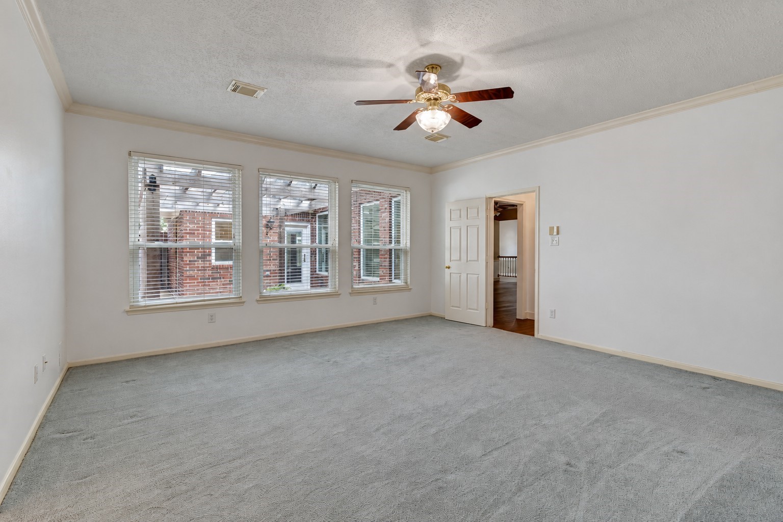 The spacious primary bedroom offers plenty of room for a king-size bed and additional furnishings. Three large windows bring in natural light, while soft carpeting, neutral walls, crown molding, and a ceiling fan create a comfortable retreat.