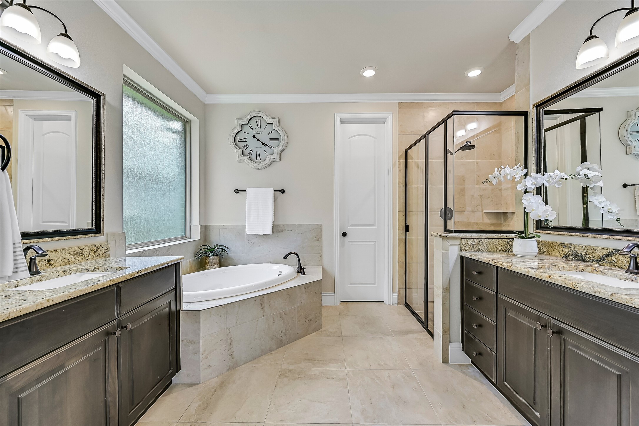 The spa-inspired primary bath offers separate granite-topped vanities with framed mirrors, stained wood cabinetry, a relaxing corner soaking tub beneath a privacy glass window, and an oversized glass-enclosed shower with tile surround and built-in shaving ledge.