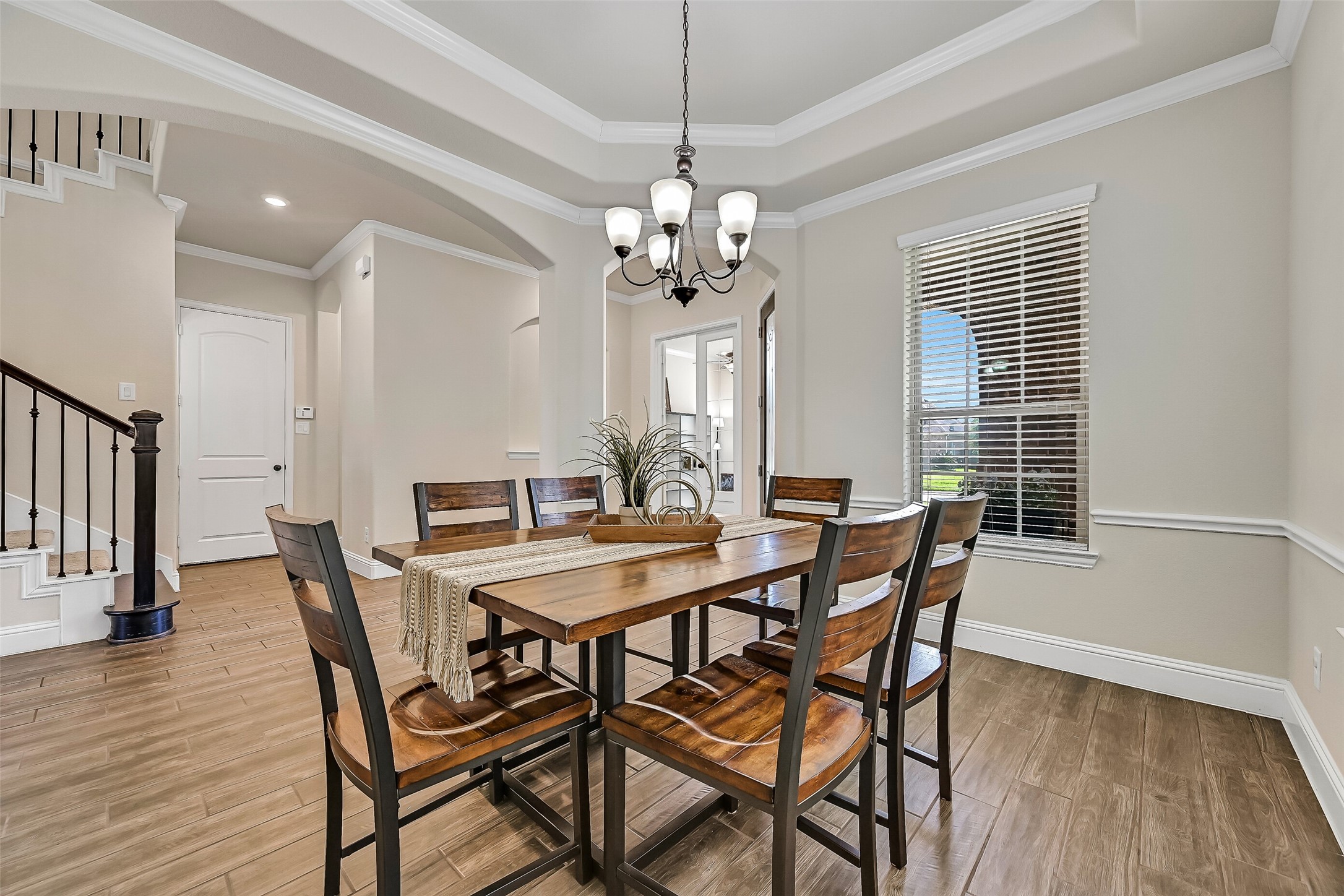 Whether hosting holiday dinners or intimate gatherings, the formal dining room provides an elegant setting with a tray ceiling, crown molding, wood-look tile flooring, abundant natural light from front and side-facing windows, and a timeless iron chandelier.
