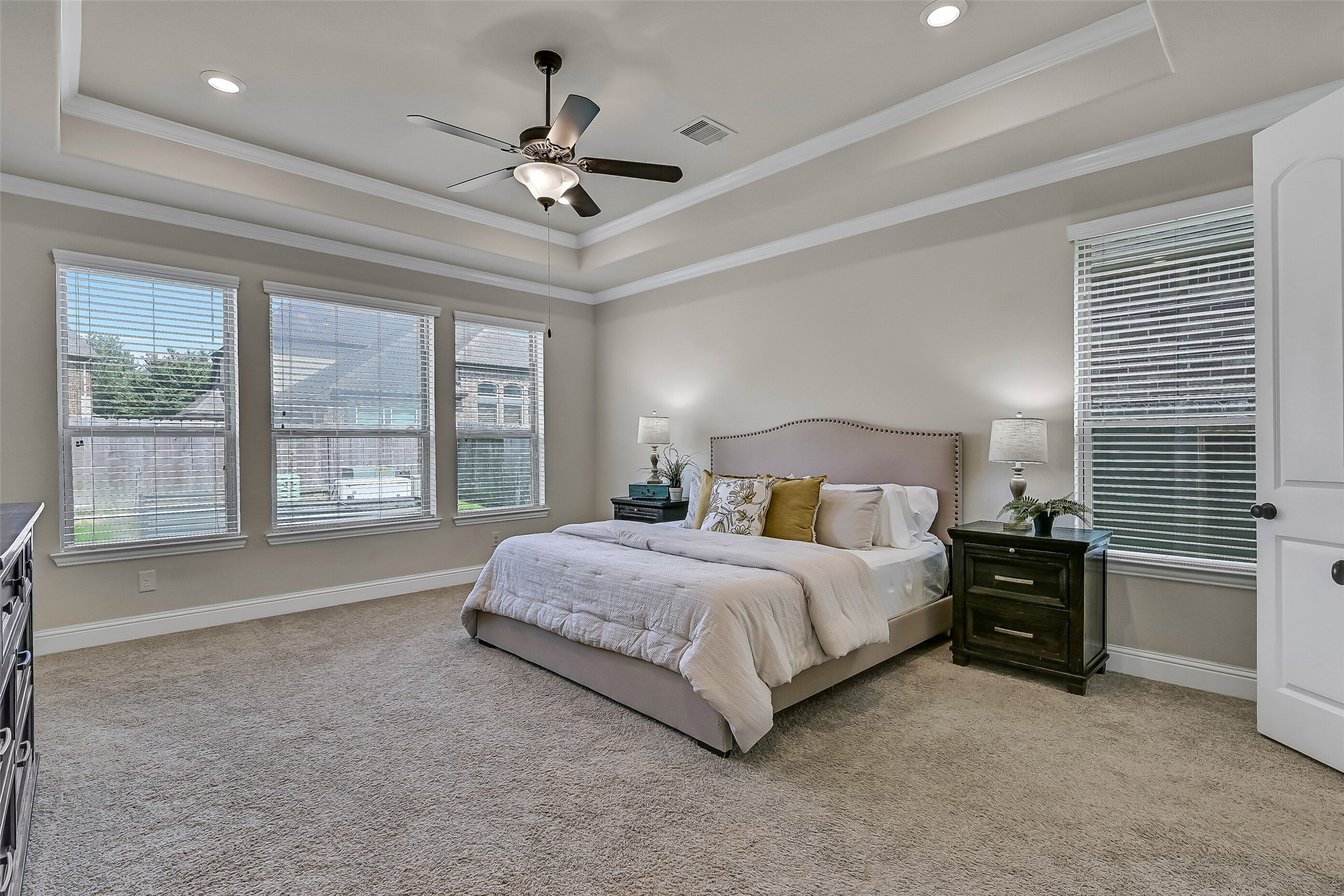 Privately tucked away on the first floor, the spacious primary suite features an elegant tray ceiling with crown molding, plush carpeting, and a wall of rear-facing windows that bring in soft natural light. The generous layout easily accommodates large furnishings while creating a peaceful retreat at the end of the day.