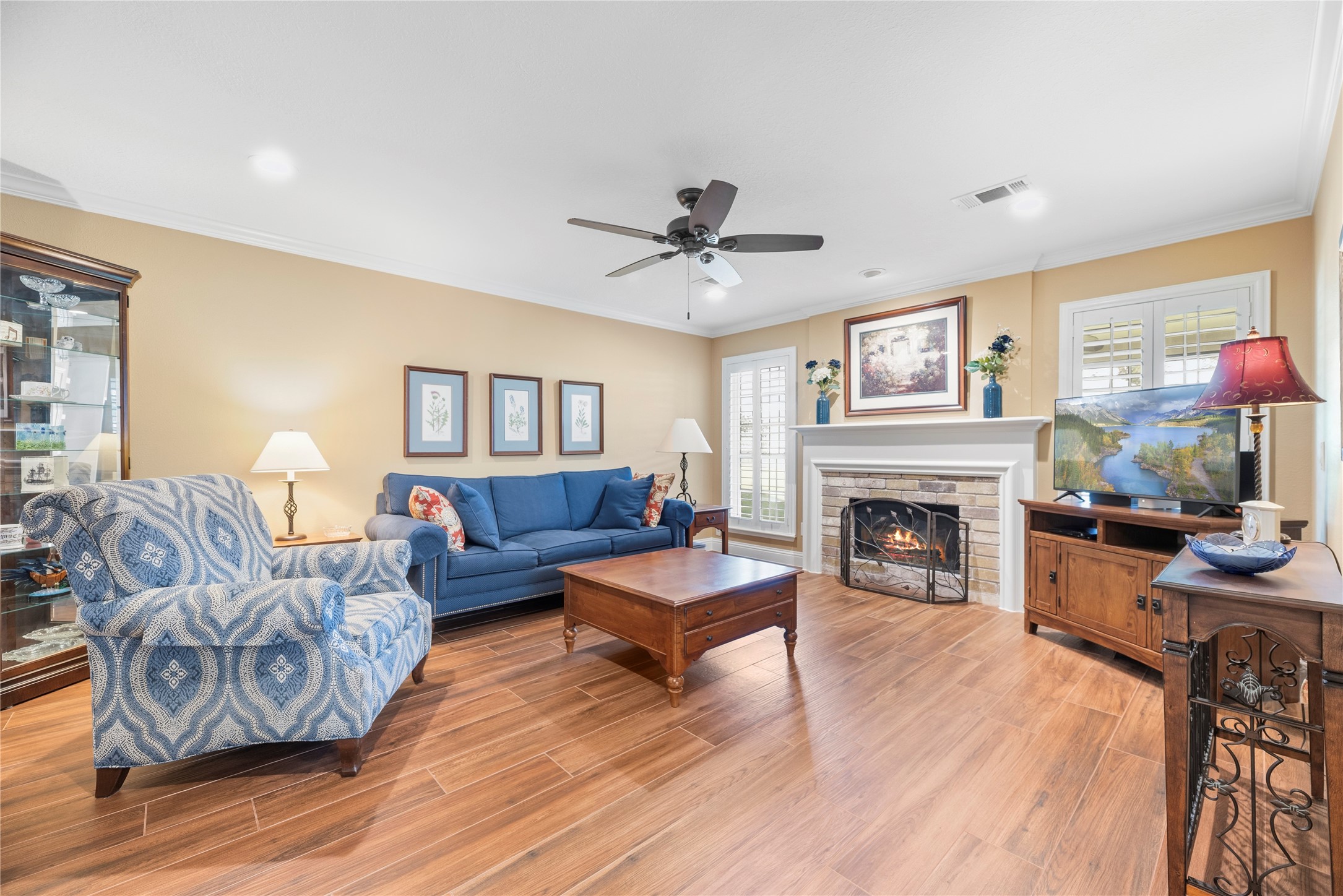 Spacious family room offers crown molding, ceiling fan, window shutters, wood-look tile flooring and a cozy fireplace with brick surround.