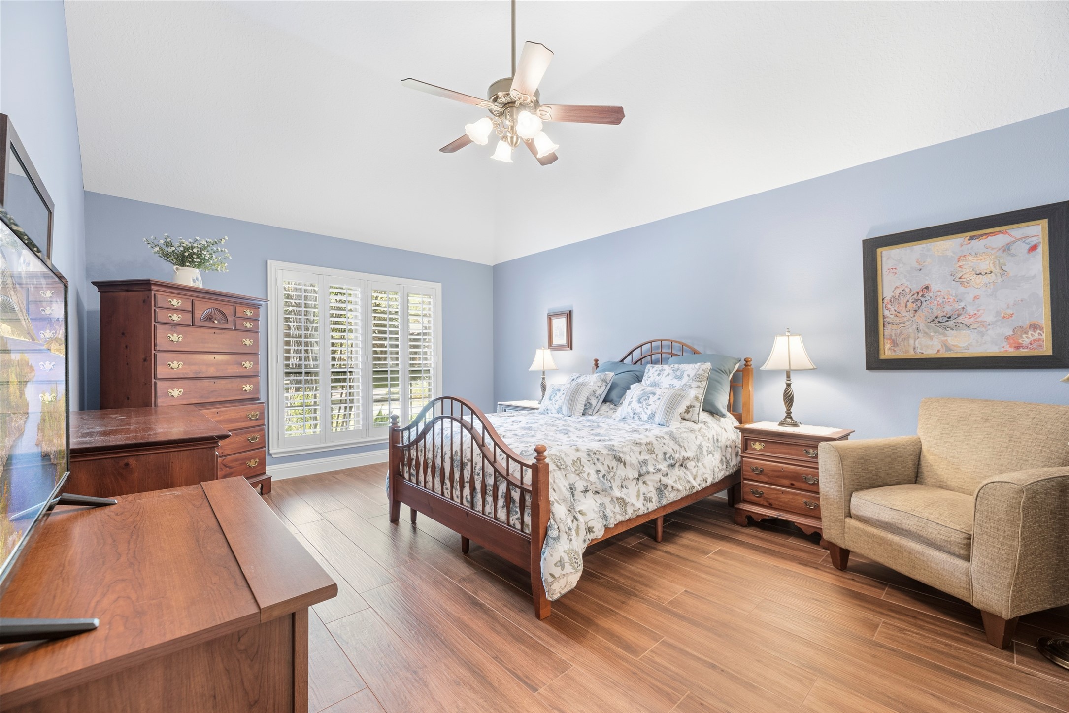 Inviting primary suite featuring a high ceiling, rich wood-look tile flooring, window shutters and plenty of natural light, with ample room for a comfortable sitting area.