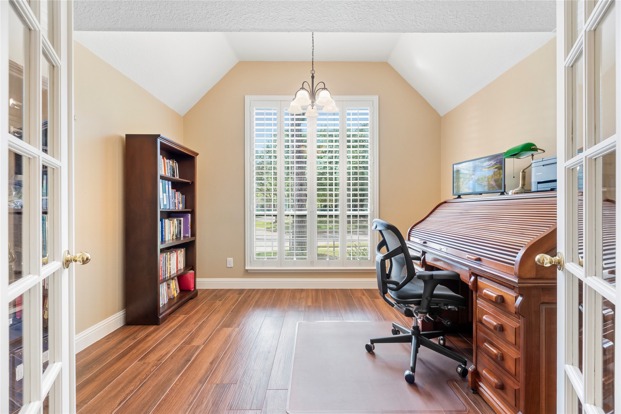 Home office features a high ceiling, window shutters, wood-look tile flooring and French doors for privacy.