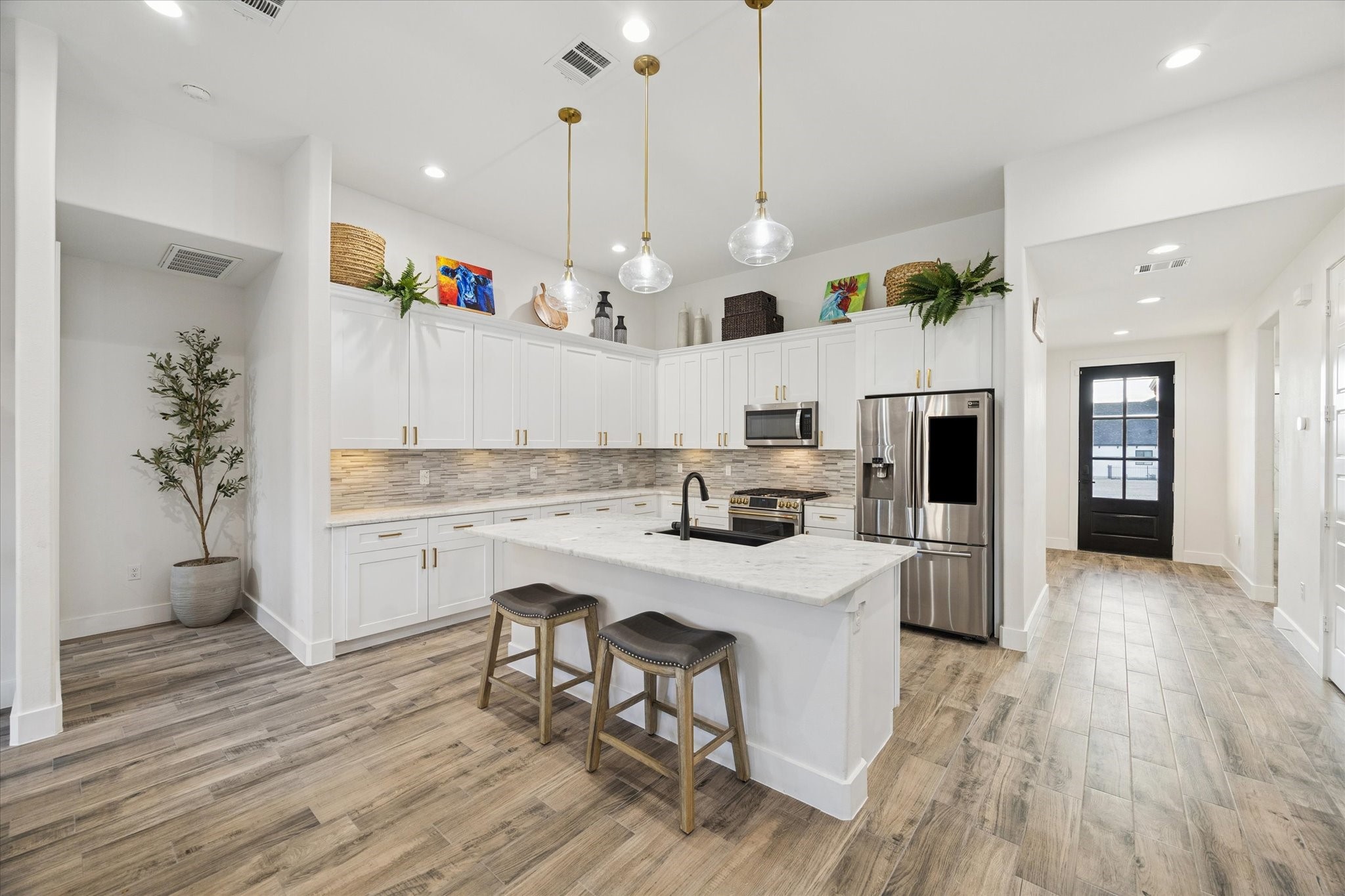 This kitchen view showcases the stunning backsplash and under-cabinet lighting, adding refined elegance to the space. The LED and pendant lights create a beautifully layered ambiance, perfectly complemented by abundant natural light and a soft, sophisticated color palette. A recently installed Litchen-Karran Farmhouse apron-front sink (34" x 21.25", single bowl, quartz) adds both style and functionality, enhancing the kitchens luxurious and inviting atmosphere.