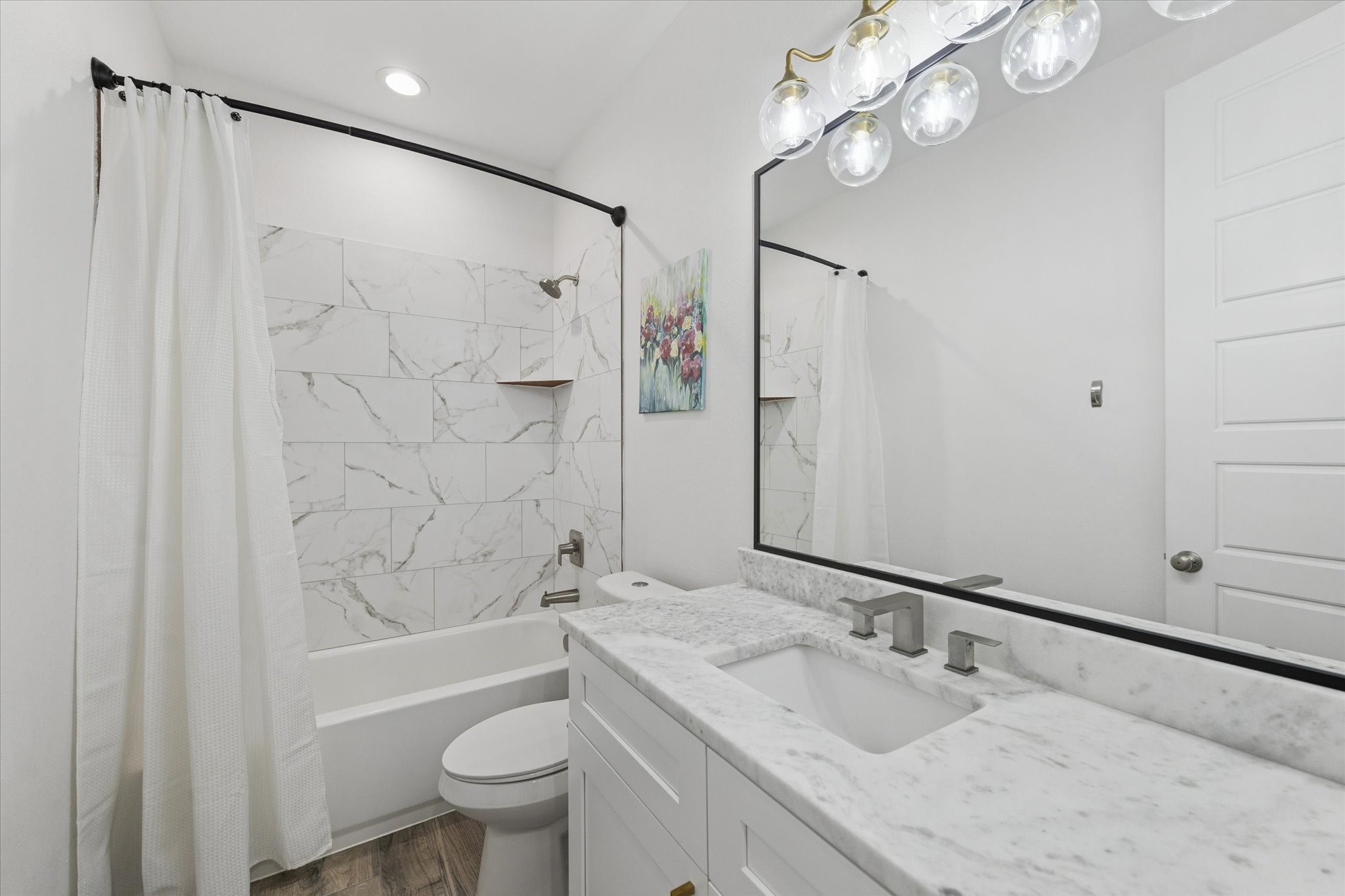 Every detail in this home reflects high-end quality, as seen in this secondary bathroom. The marble countertops, clear tile in the bathtub area, and custom mirror with dark edging create a harmonious and sophisticated look. Recently installed dark faucets and the brass LED light fixture enhance the space, making it both functional and stylish. This serene bathroom offers a calm and inviting retreat for family members or guests.