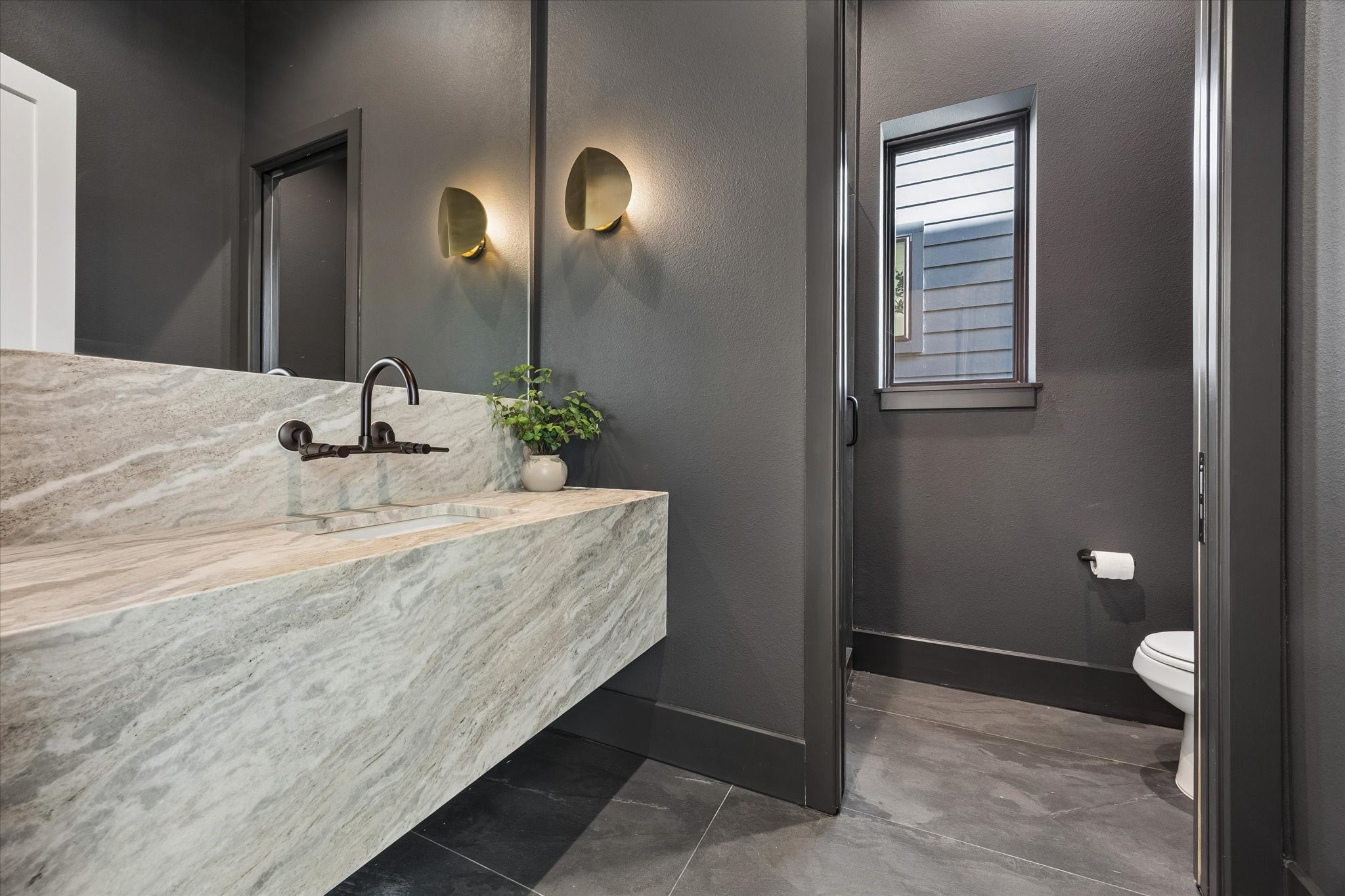 Moody and memorable, the powder bath pairs deep charcoal walls with a dramatic stone vanity, integrated sink and wall mounted faucet. Sculptural brass sconces bring warmth to the richly layered palette.