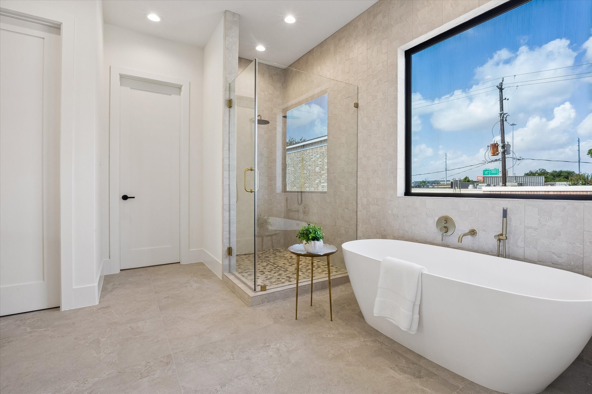 Freestanding soaking tub sits beneath a large picture window, while the adjacent walk in shower features floor to ceiling stone tile, dual shower heads, and warm brass fixtures.