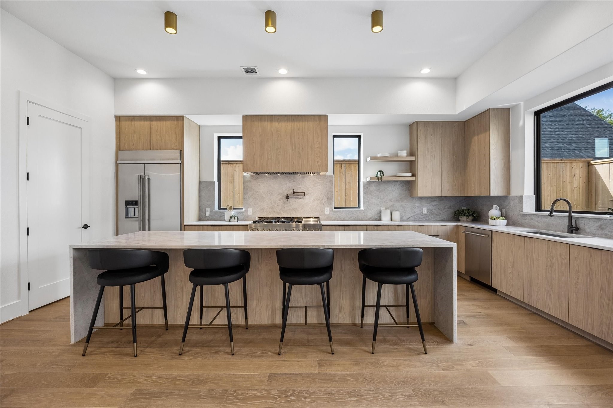 Natural oak cabinetry gives the kitchen a warm, tailored look, complemented by a stone backsplash that carries seamlessly behind the range. An expansive waterfall island offers seating for four, while slim windows and curated open shelving keep the design light and architectural.