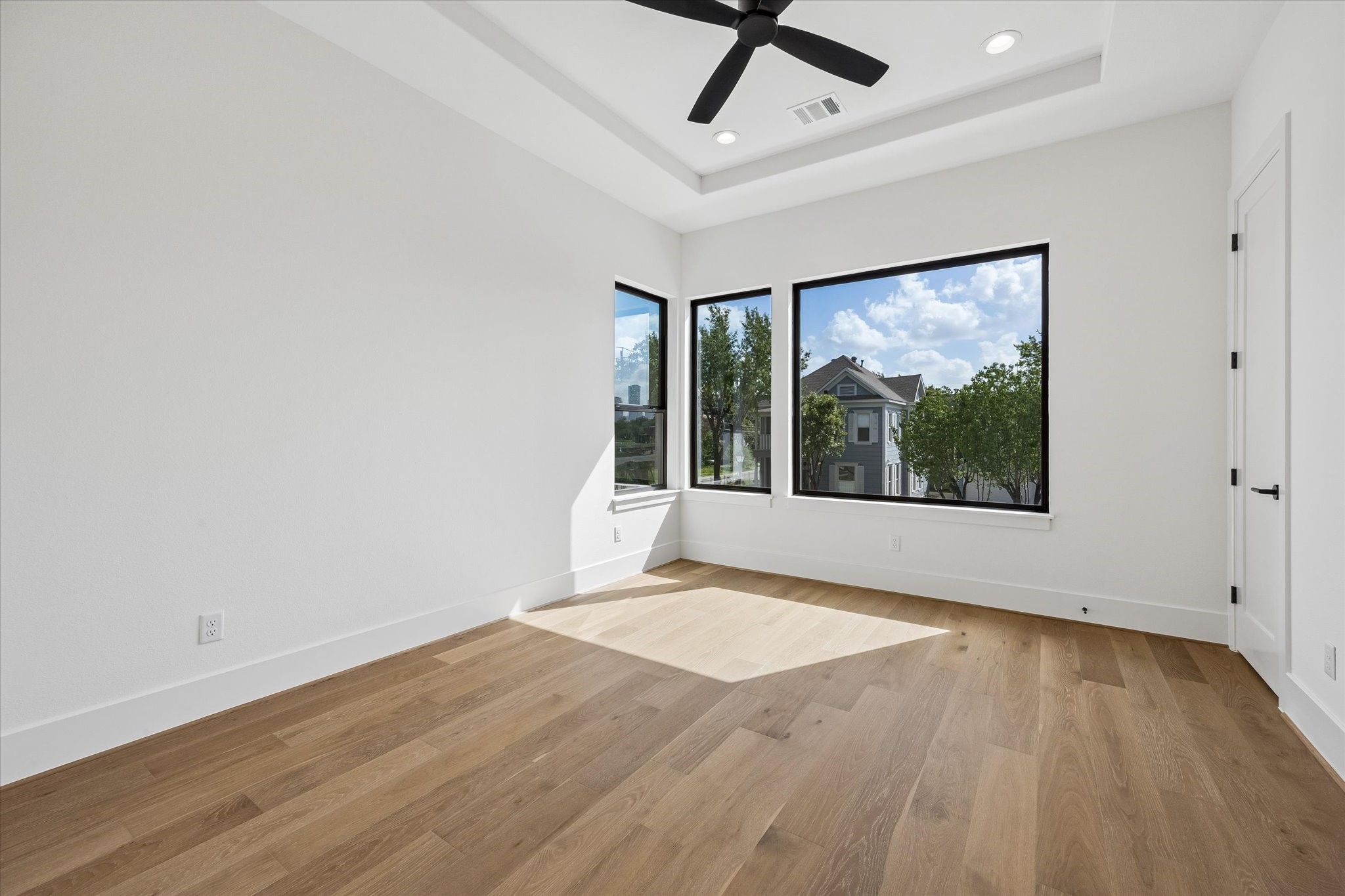 Natural light pours into the secondary bedroom through oversized black framed windows, complemented by warm hardwood floors and a tray ceiling that adds architectural dimension.