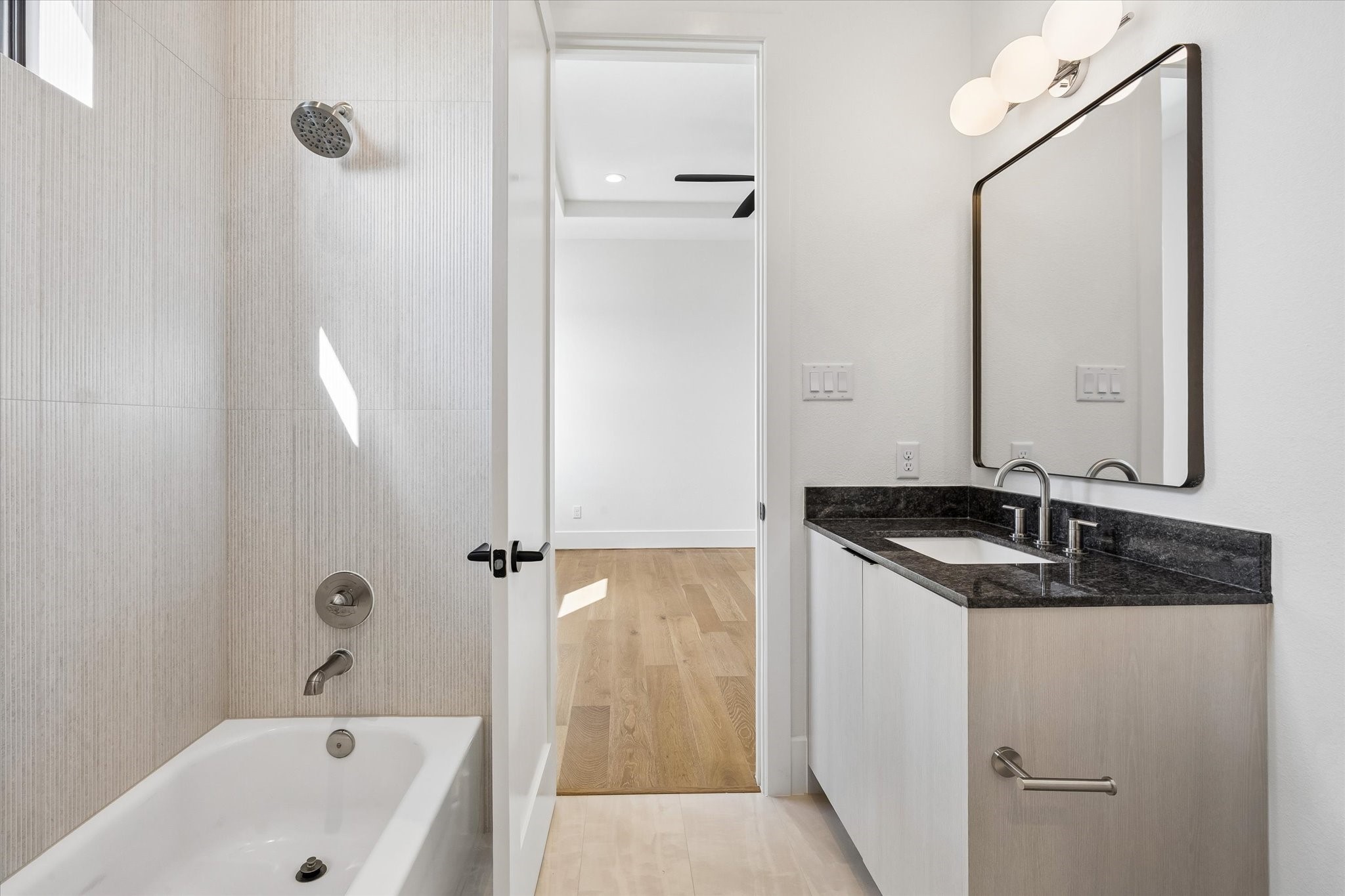 Contemporary finishes continue into the secondary ensuite, pairing a dark stone countertop with light wood cabinetry and softly textured tile surrounding the tub and shower.
