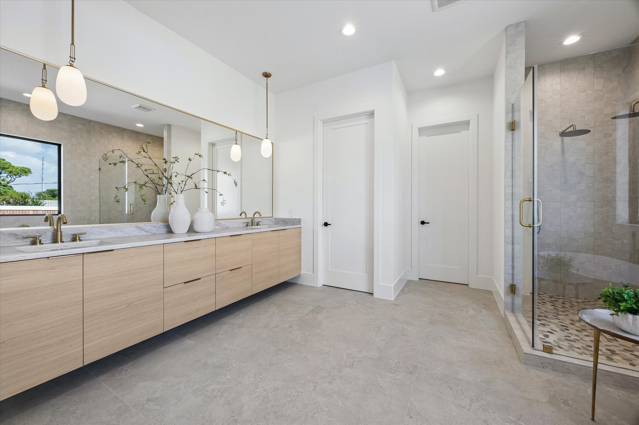 An expansive floating vanity stretches beneath an oversized mirror, pairing warm wood cabinetry with stone counters and understated brass fixtures. The glass enclosed shower continues the neutral stone palette for a cohesive, spa inspired finish.