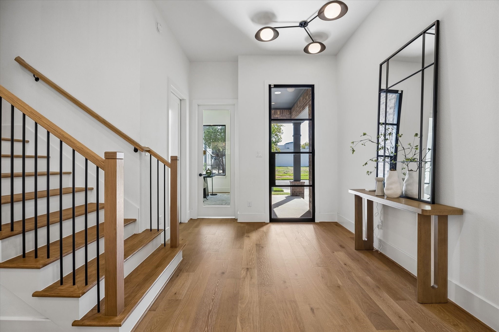 Warm wood flooring and a streamlined staircase with black metal balusters bring contrast and character to the light filled entry. A modern statement fixture and tall glass front door introduce the homes clean, contemporary design from the start. Warm wood flooring and a streamlined staircase with black metal balusters bring contrast and character to the light filled entry. A modern statement fixture and tall glass front door introduce the homes clean, contemporary design from the start.