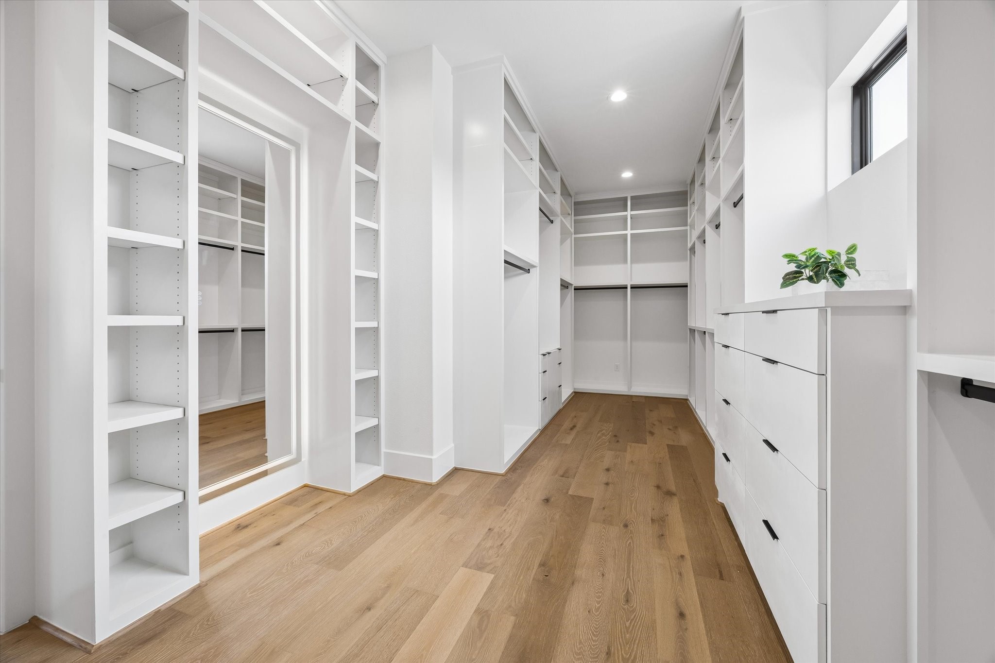 Custom built ins transform the primary walk in closet with extensive shelving, hanging space, integrated drawers, and a full length mirror. A window introduces natural light while hardwood flooring carries through from the primary suite.