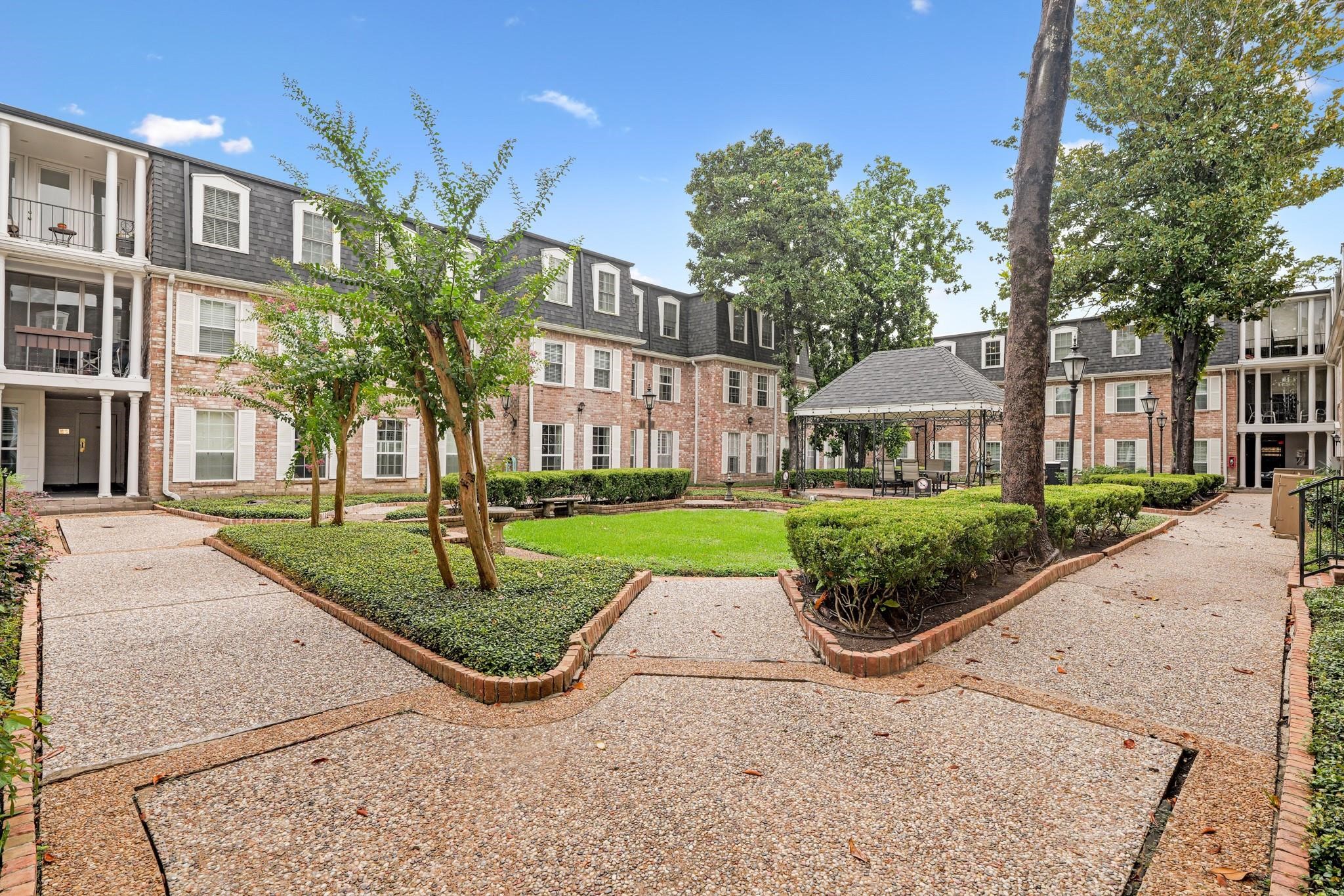 Charming courtyard view of a classic brick apartment complex featuring a lush central lawn, mature trees, and a cozy gazebo, offering a serene and inviting atmosphere.